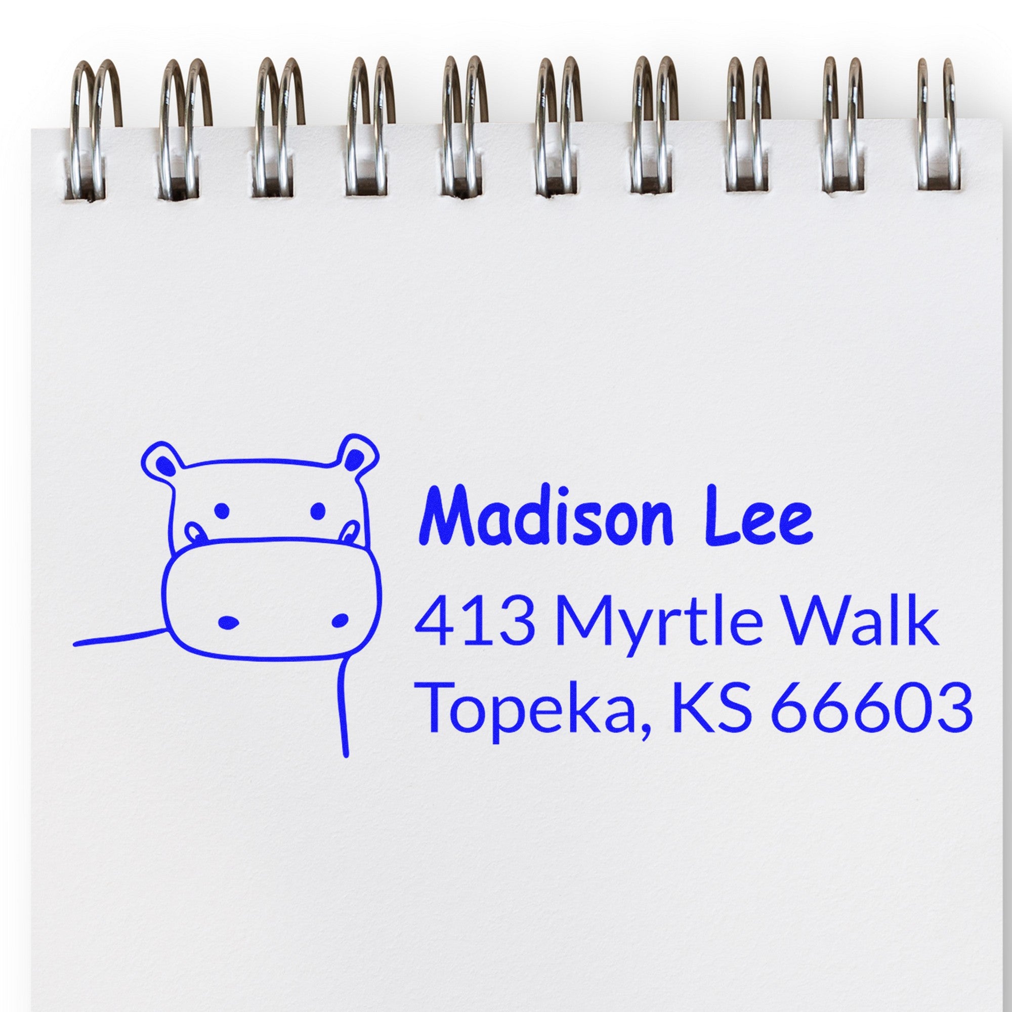 PSI Happy Hippo Handmade New Address Pre-Inked Stamp on a notepad, featuring a customizable home stamp design with a cute hippo illustration and the address "413 Myrtle Walk, Topeka, KS 66603" in blue ink.