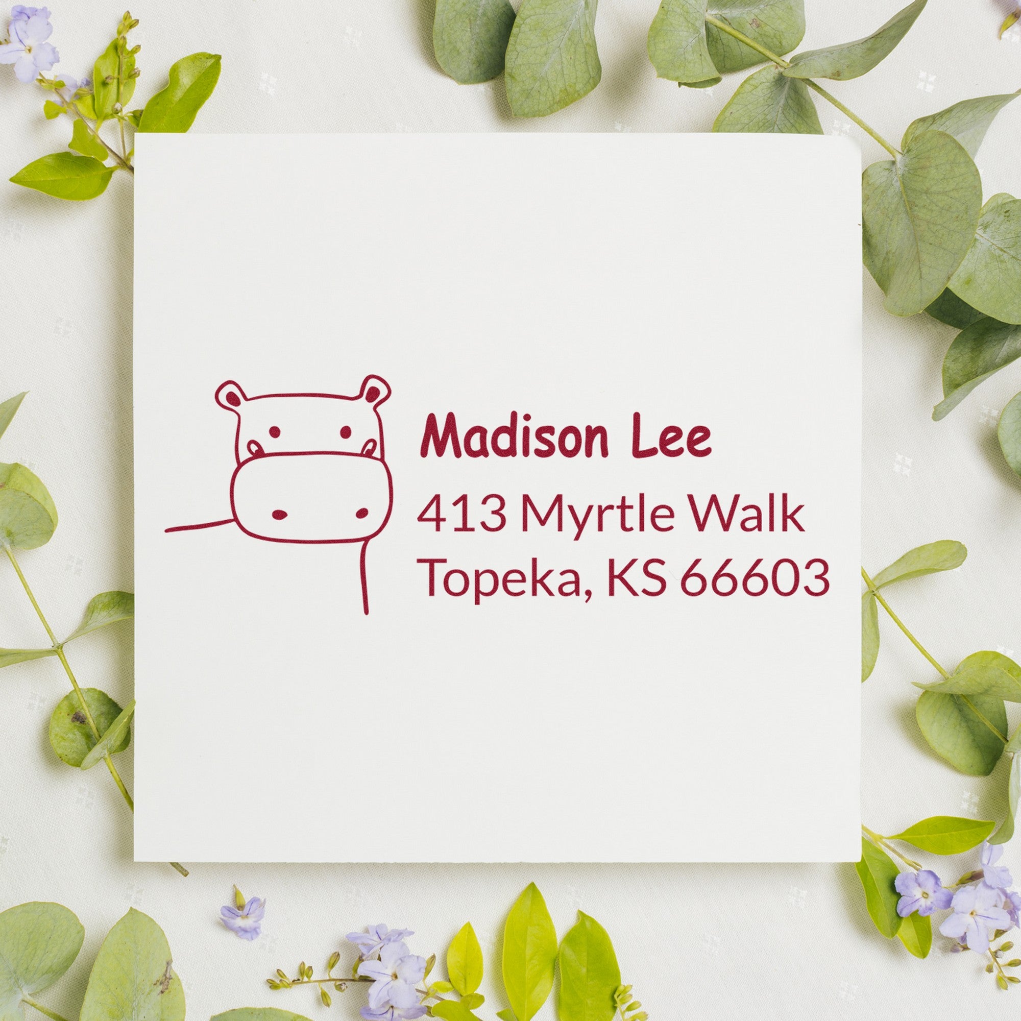 PSI Happy Hippo Handmade New Address Pre-Inked Stamp on a white card with green leaves and purple flowers. The Customizable New Address Stamper displays "Madison Lee, 413 Myrtle Walk, Topeka, KS 66603" in red ink.