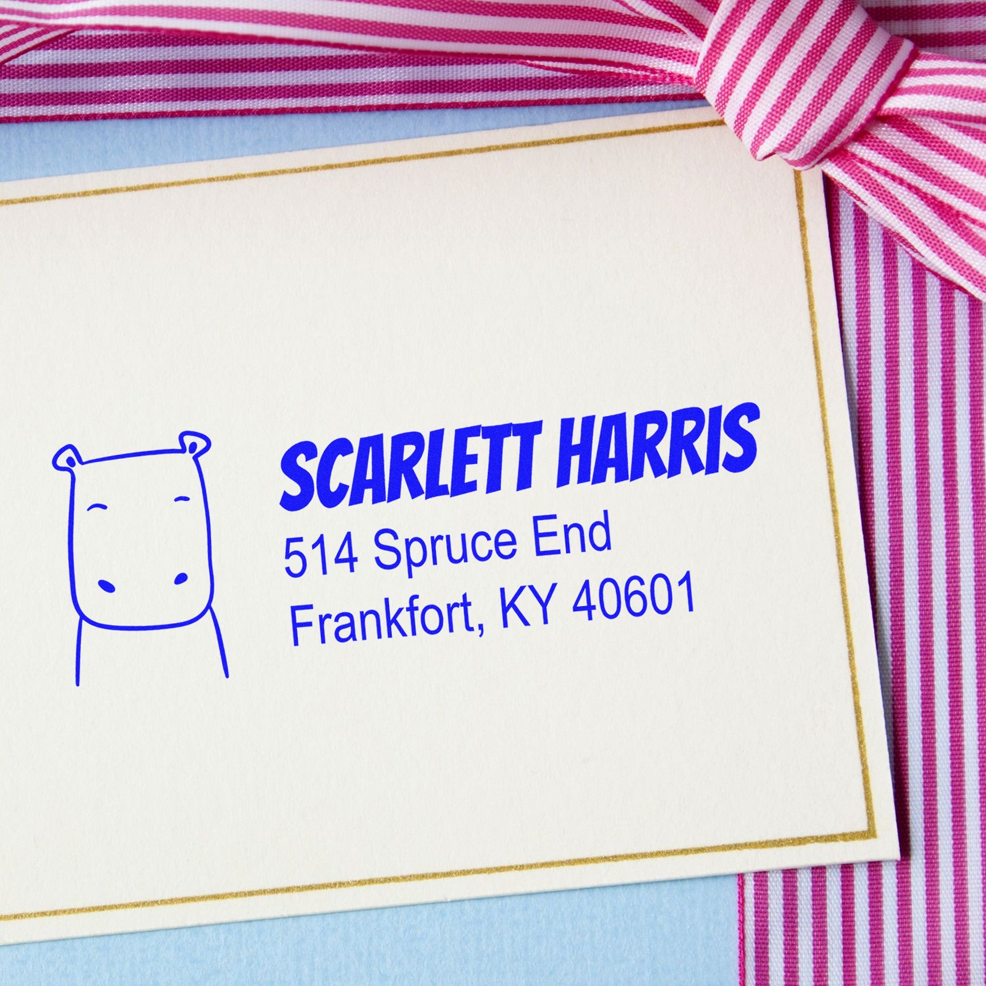 Image of a card with a Wood Handle Hopeful Hippo Custom Mail Rubber Stamp design, featuring a hippo illustration and text: "Scarlett Harris, 514 Spruce End, Frankfort, KY 40601." This Custom New Address Stamp is placed on a card with a pink striped ribbon.