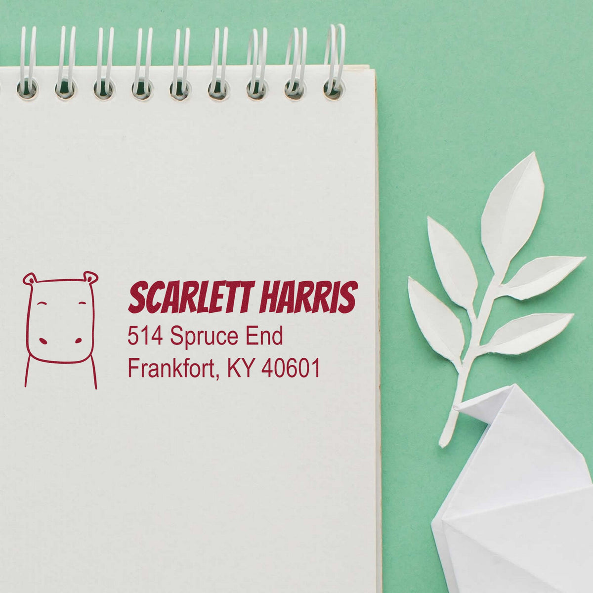 Slim Pre-Inked Hopeful Hippo Customized Return Address Stamp on a notepad with a hippo design. Text reads "Scarlett Harris, 514 Spruce End, Frankfort, KY 40601." Personalizable New Address Stamper shown with paper leaves on a green background.