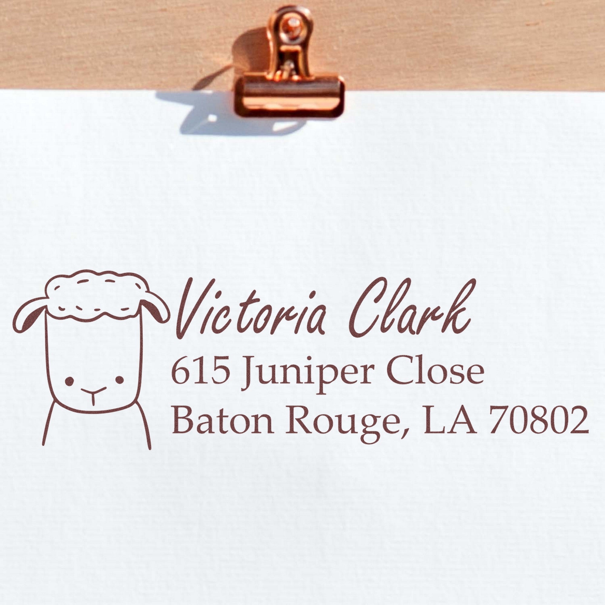 Fluffy Sheep Customizable Mail Address Self-Inking Stamp featuring a cute sheep design and the text "Victoria Clark, 615 Juniper Close, Baton Rouge, LA 70802." This personalizable address stamp is shown on white paper clipped to a wooden board.