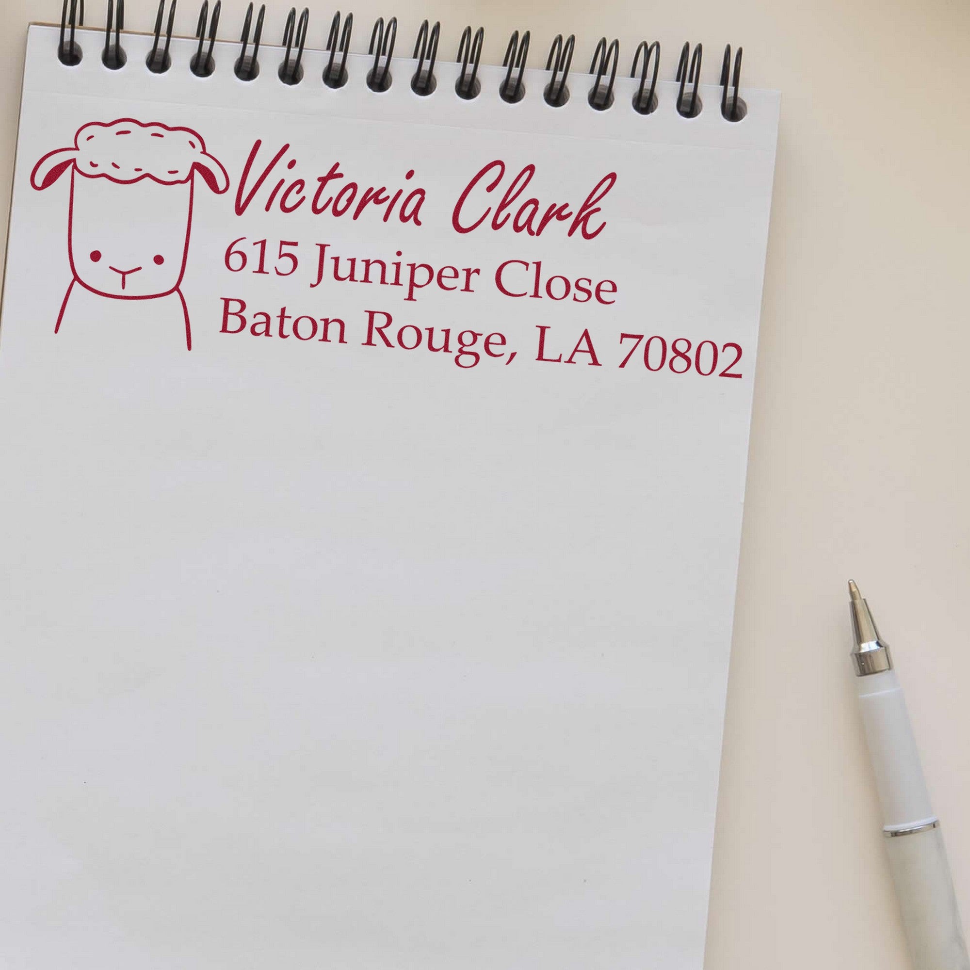 Wood Handle Fluffy Sheep Custom Home Address Rubber Stamp on a notepad, displaying "Victoria Clark, 615 Juniper Close, Baton Rouge, LA 70802" in red ink. Customized Mailing Stamper design features a cute sheep illustration. Pen placed beside the notepad.