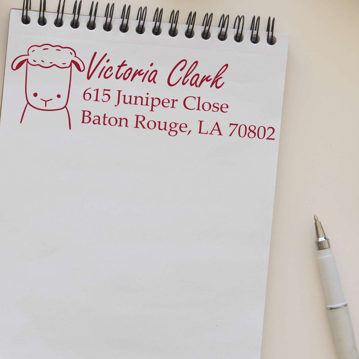 Slim Pre-Inked Fluffy Sheep Customized Name and Address Stamp on a notepad, displaying "Victoria Clark, 615 Juniper Close, Baton Rouge, LA 70802" in red ink. A pen lies beside the notepad. Ideal for a custom mailing stamper.