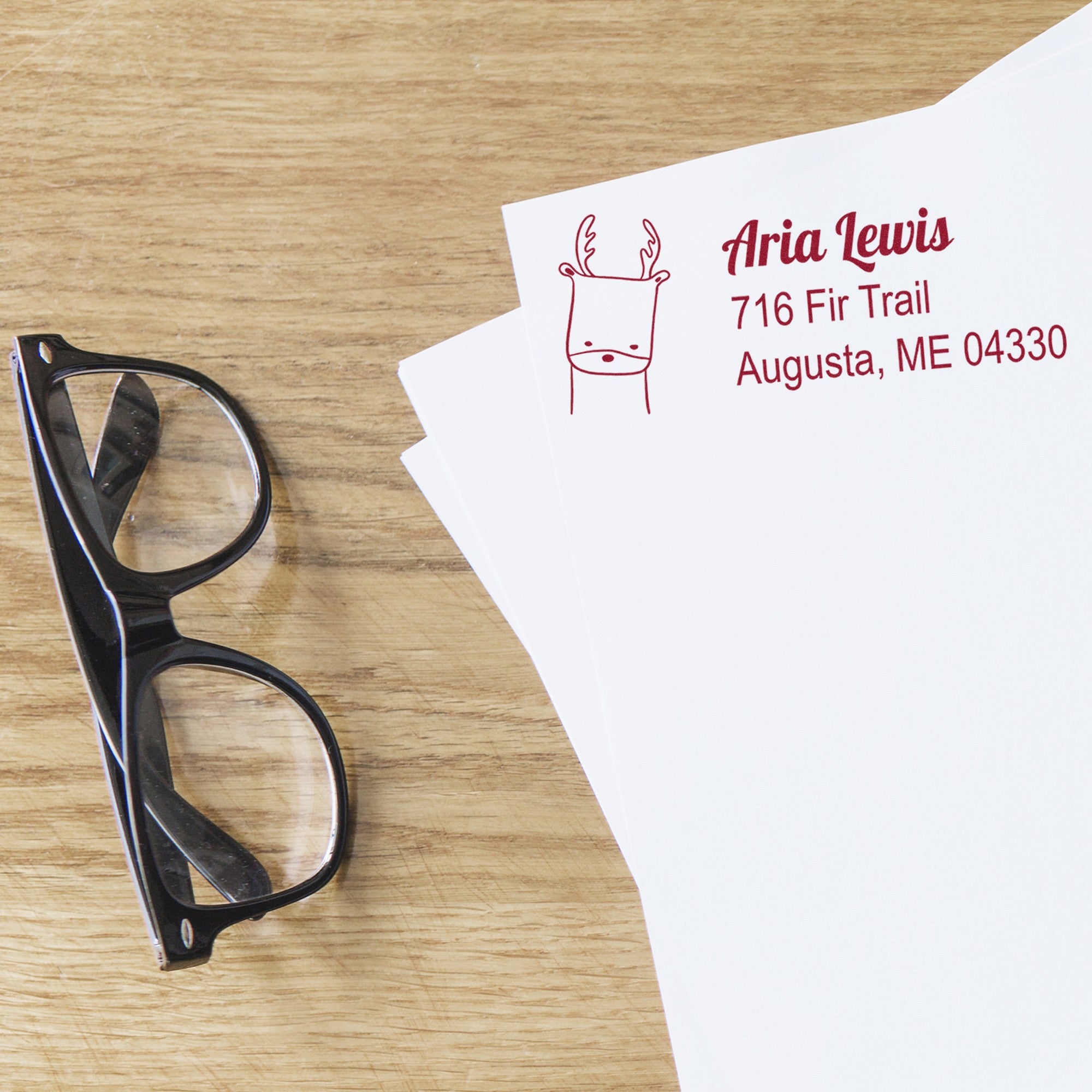 Slim Pre-Inked Darling Deer Customized Mailing Stamp on white envelopes with red ink, featuring a deer illustration and custom-made address stamp for Aria Lewis, 716 Fir Trail, Augusta, ME 04330. Black glasses rest nearby on a wooden surface.