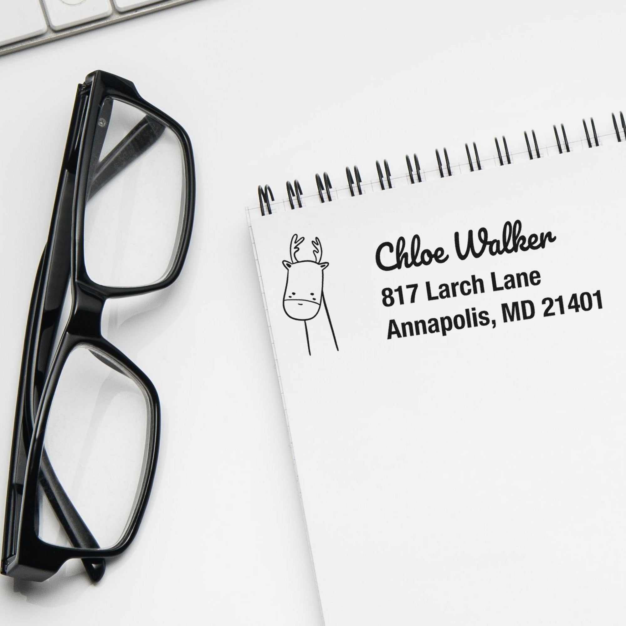 Wood Handle Regal Reindeer Custom Mailing Address Rubber Stamp on a notepad with customizable address stamp featuring a reindeer design. Nearby are black-framed glasses and a keyboard, creating a stylish and personalized stationery setup.