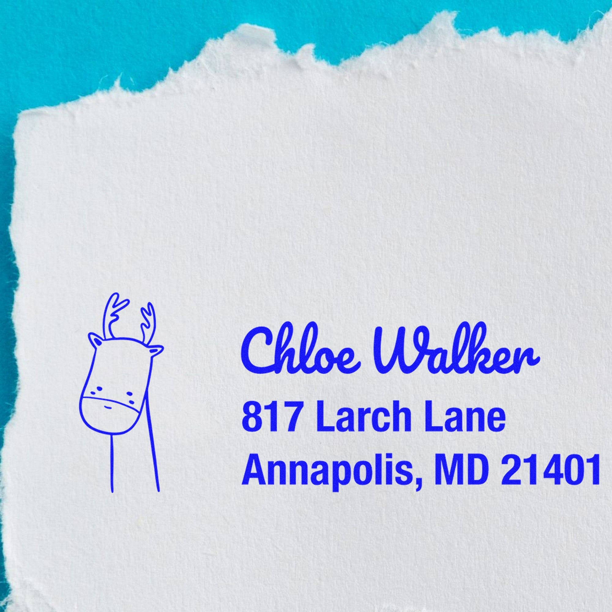 A PSI Regal Reindeer Customize Return Address Pre-Inked Stamp impression on torn white paper against a blue background. The stamp features a reindeer illustration and reads Chloe Walker, 817 Larch Lane, Annapolis, MD 21401. It's a Customizable Home Stamp.