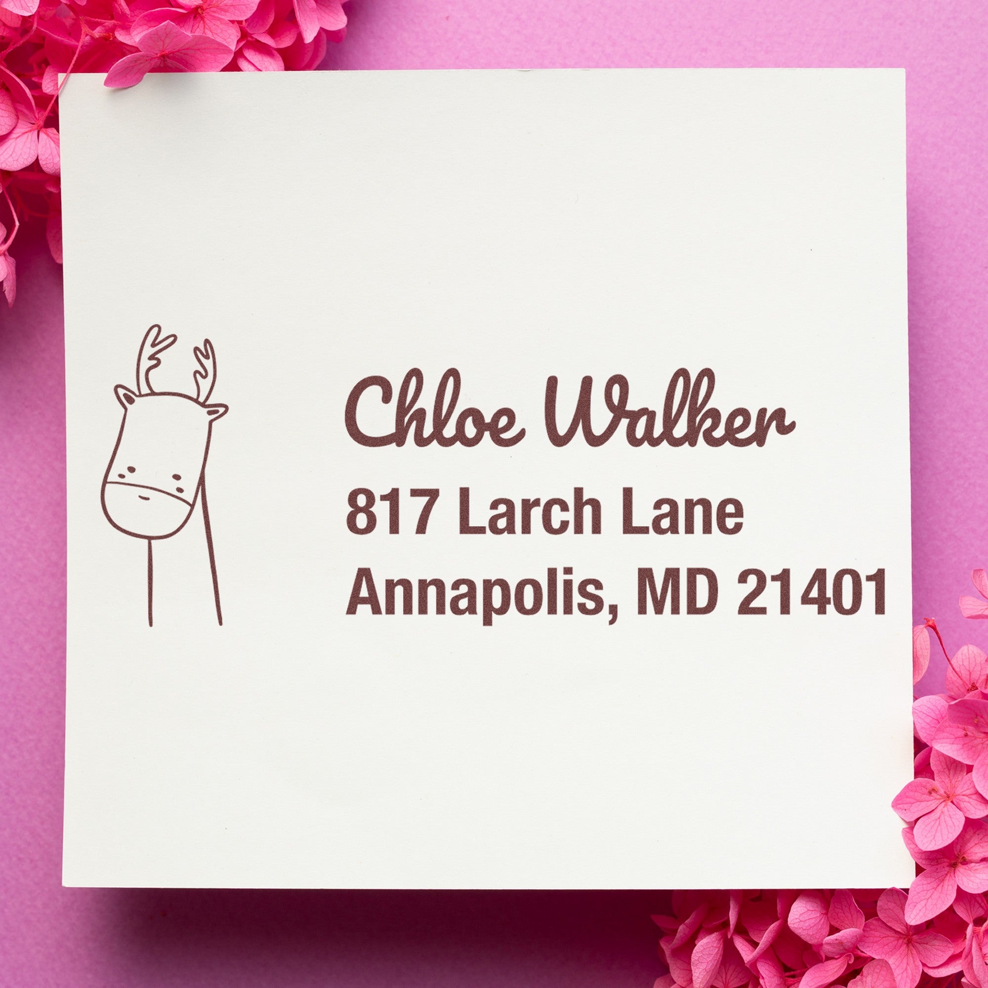 A Wood Handle Regal Reindeer Custom Mailing Address Rubber Stamp is used on a white card with pink flowers. The stamp features a reindeer design and customizable home stamp details, displaying a name and address in brown ink.