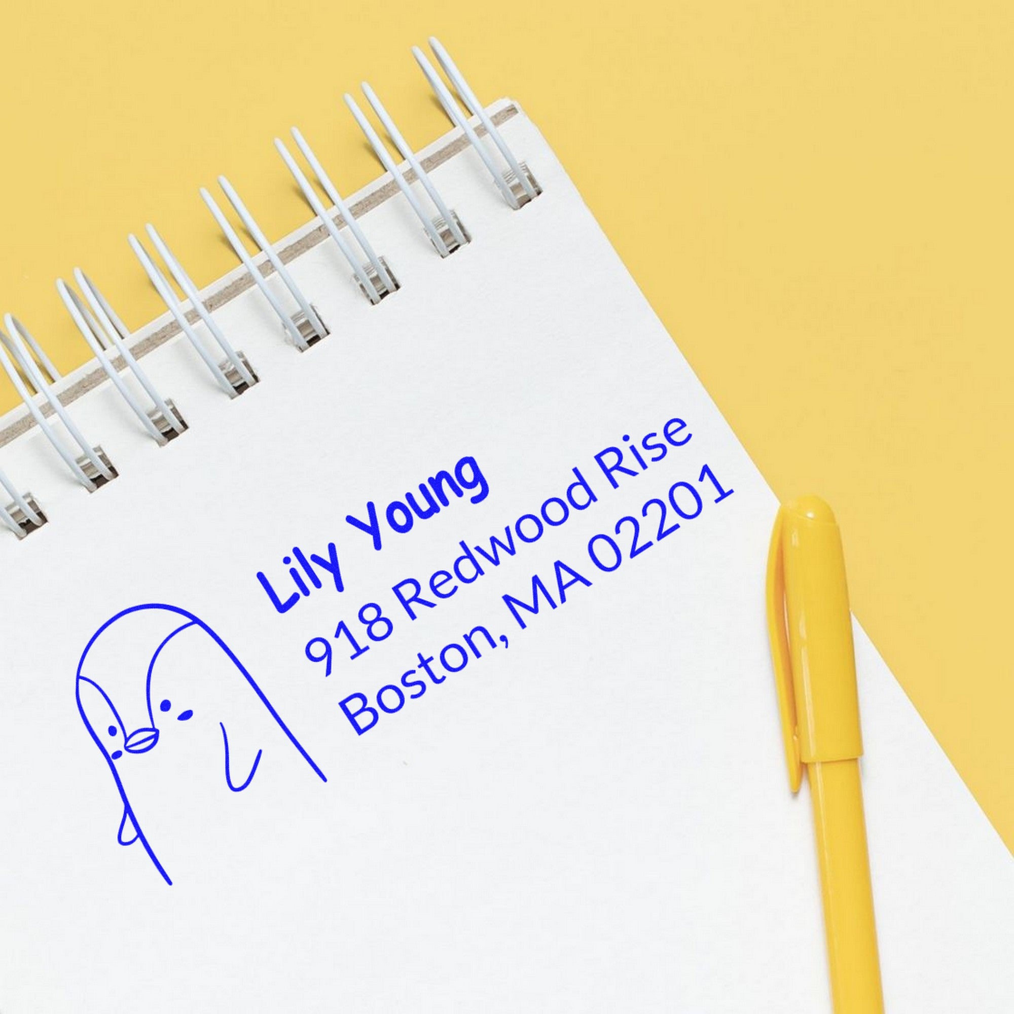 Slim Pre-Inked Perky Penguin Customized Home Address Stamp on a notepad with a yellow pen. The Customizable Mailing Stamper displays a penguin graphic and address: Lily Young, 918 Redwood Rise, Boston, MA 02201, in blue ink on white paper.