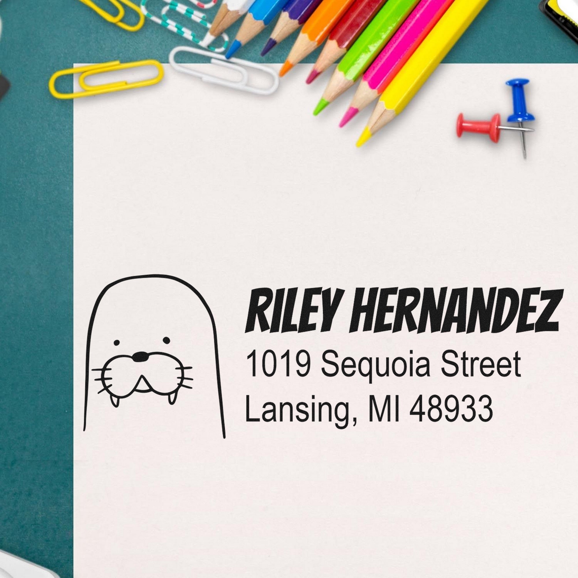 Wood Handle Wacky Walrus Custom New Address Rubber Stamp on paper with "Riley Hernandez, 1019 Sequoia Street, Lansing, MI 48933." Surrounded by colorful pencils and paperclips. Personalizable Home Stamper for creative address labeling.