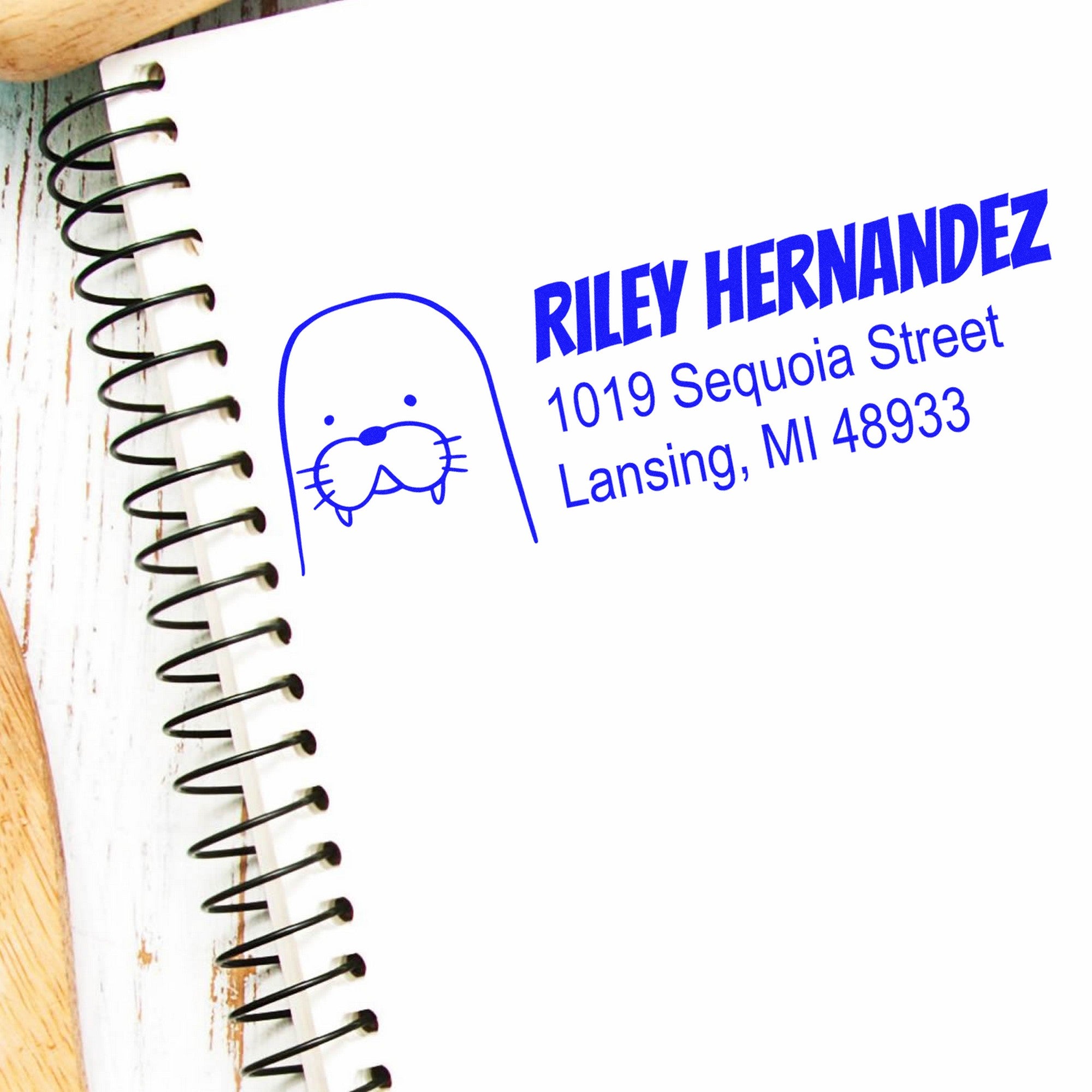 Image of a notebook with a blue stamped design featuring a walrus and text. The stamp reads "Riley Hernandez, 1019 Sequoia Street, Lansing, MI 48933." Keywords: Wood Handle Wacky Walrus Custom New Address Rubber Stamp, Personalizable Mailing Stamp.