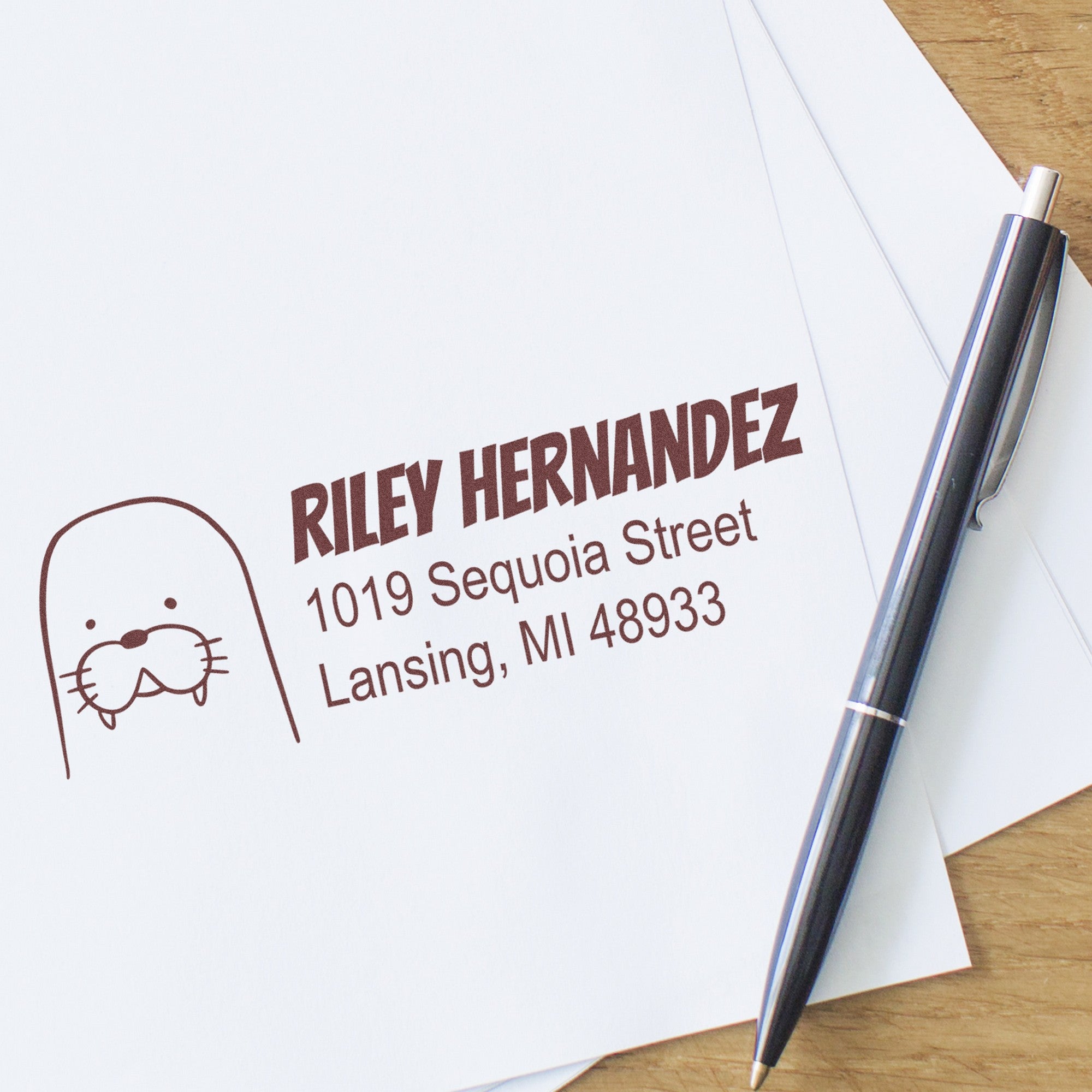 PSI Wacky Walrus Customize Mailing Pre-Inked Stamp used on an envelope, featuring a walrus graphic and the text "Riley Hernandez, 1019 Sequoia Street, Lansing, MI 48933." This Personalizable Home Stamp is shown next to a pen on a wooden surface.