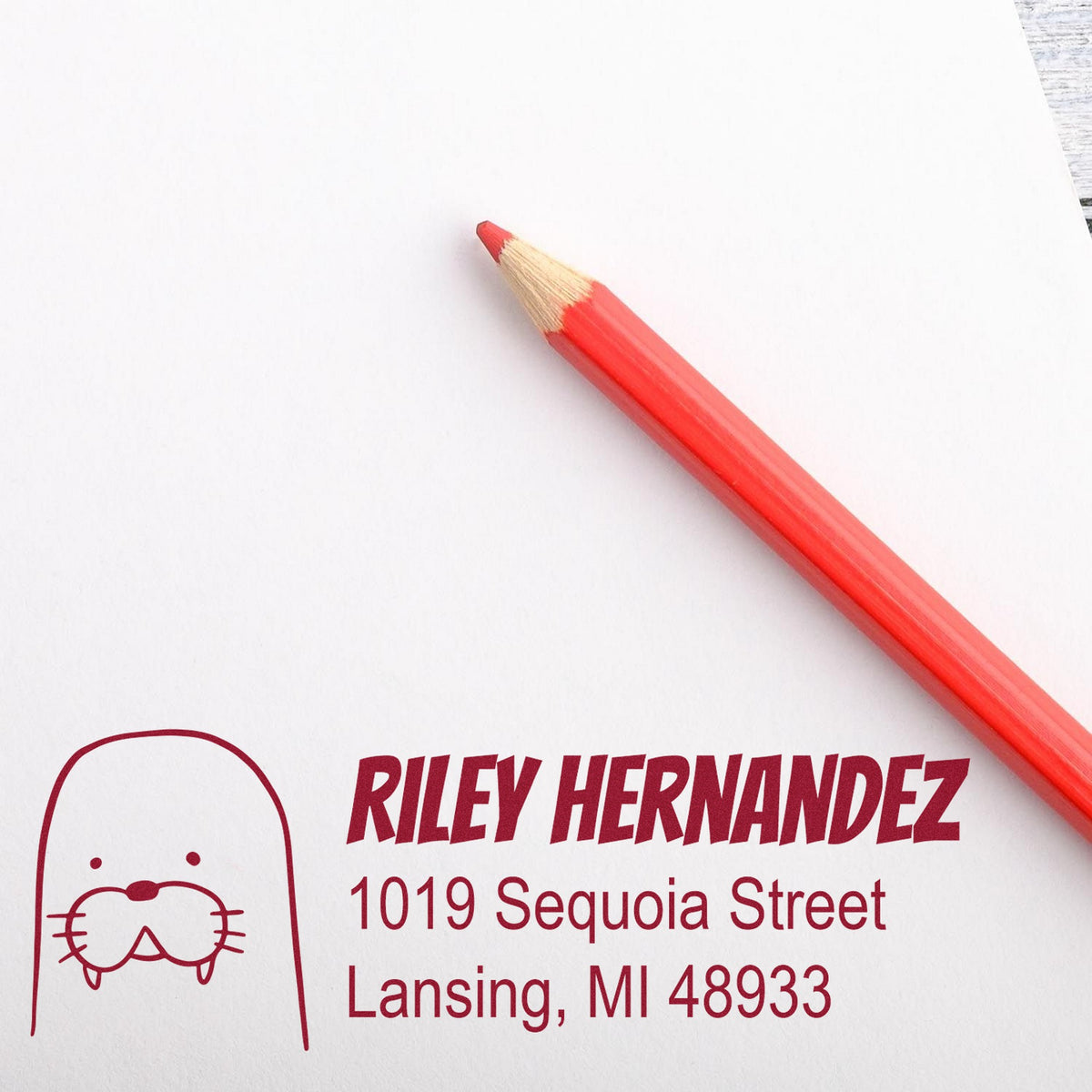 A red pencil lies on white paper stamped with a Slim Pre-Inked Wacky Walrus Customized Address Return Stamp. The stamp features a walrus design and reads "Riley Hernandez, 1019 Sequoia Street, Lansing, MI 48933," showcasing a Custom-Made New Address Stamp.