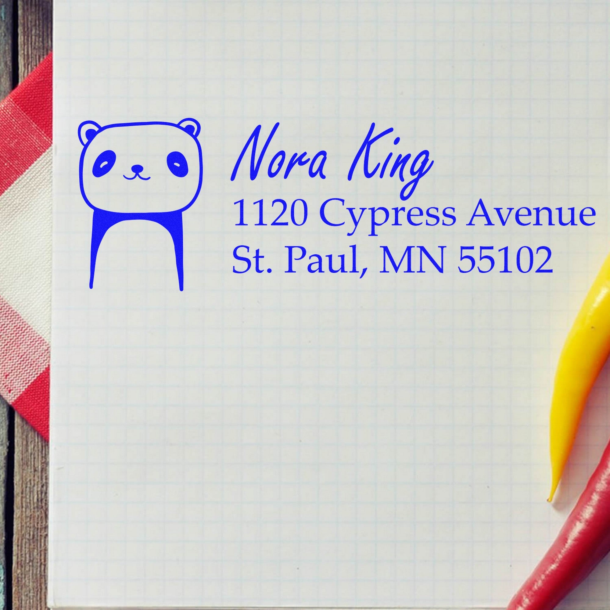 Image of a customized mailing stamper featuring a playful panda design with a wood handle. The stamp displays a home address for envelopes: "Nora King, 1120 Cypress Avenue, St. Paul, MN 55102," on a grid paper background with a red and white cloth.