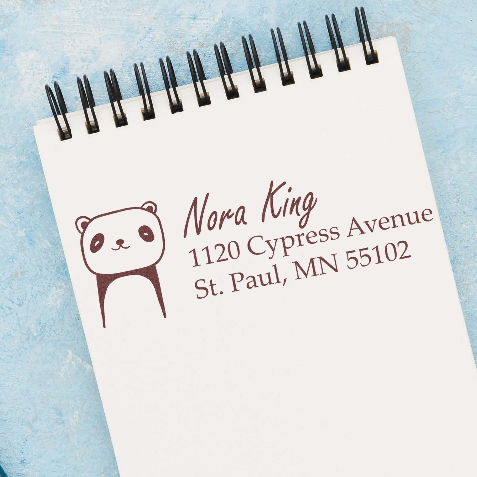Playful Panda Personalized Return Address Self-Inking Stamp on a notepad, featuring a cute panda design with the name "Nora King" and address. This Personalized Mailing Stamp adds charm to correspondence, set against a light blue textured background.