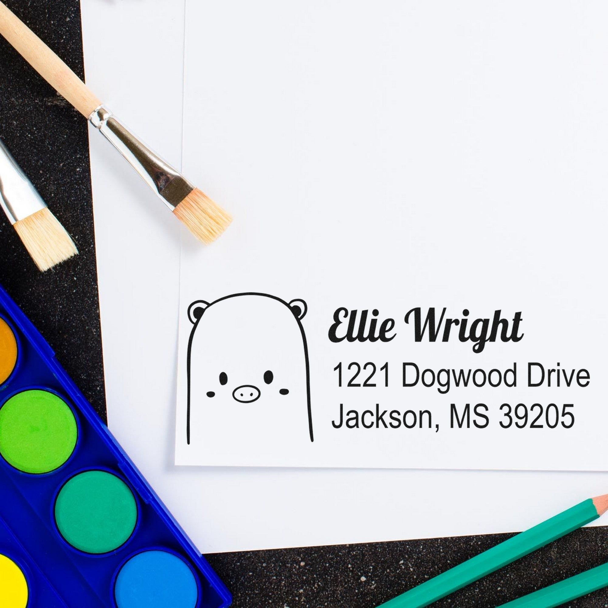 Wood handle beautiful bear custom address label rubber stamp on white paper, featuring a cute bear design. Personalizable return mail stamper with the name "Ellie Wright" and address "1221 Dogwood Drive, Jackson, MS 39205" beside paintbrushes and watercolor paints.