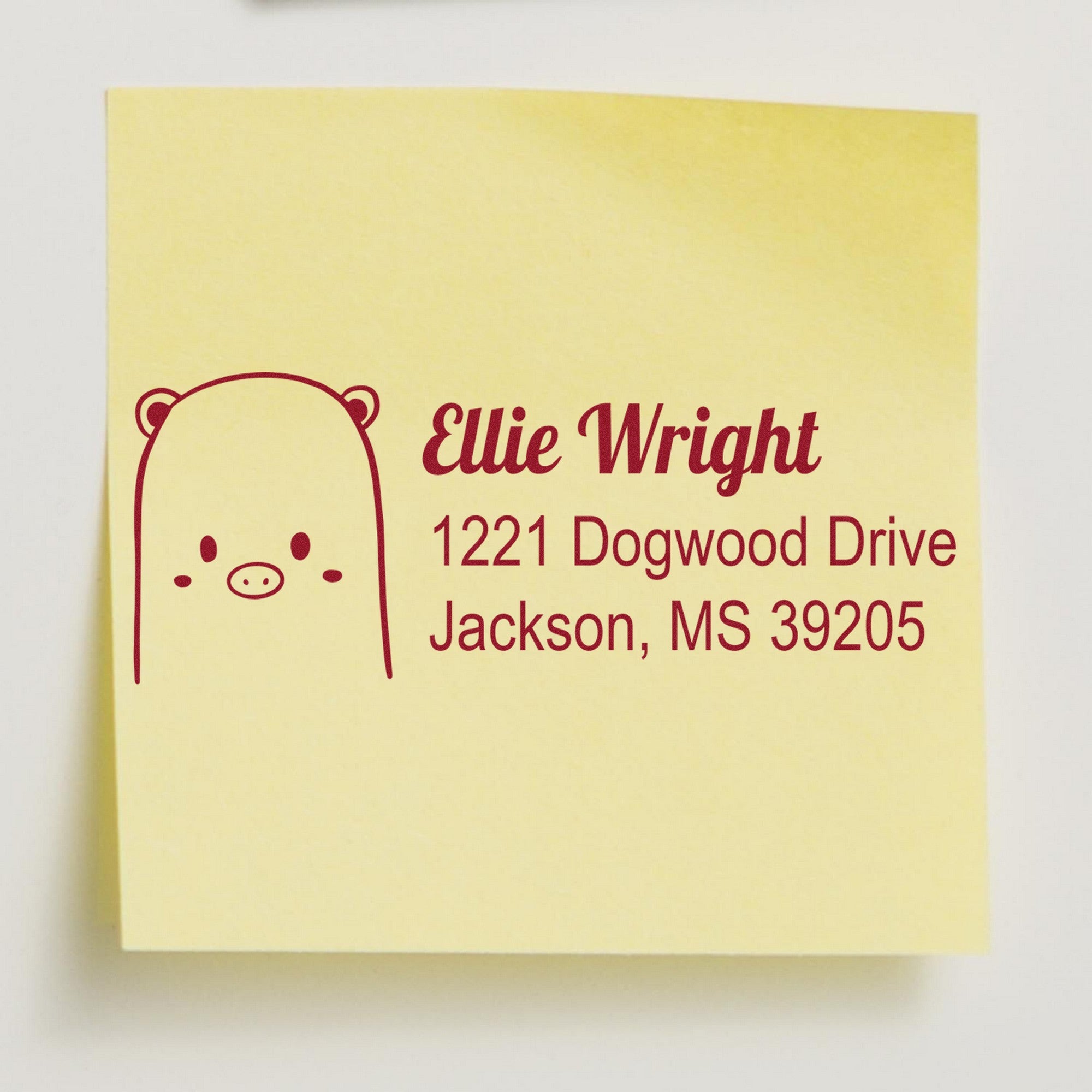 A personalized home stamp with a wood handle, featuring a beautiful bear design, is used to create a custom address label on a yellow sticky note. The label reads Ellie Wright, 1221 Dogwood Drive, Jackson, MS 39205.