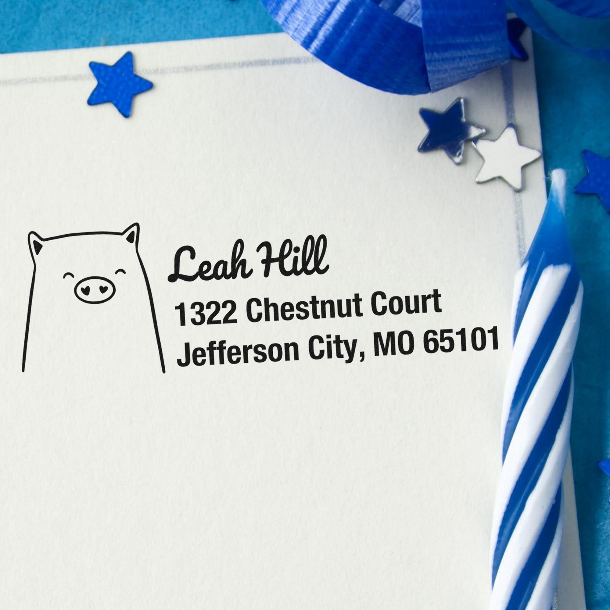 PSI Cheery Pig Customize Address Return Pre-Inked Stamp on a card with "Leah Hill, 1322 Chestnut Court, Jefferson City, MO 65101." Blue stars and a striped candle are nearby. Customized New Address Stamper design with a pig illustration.
