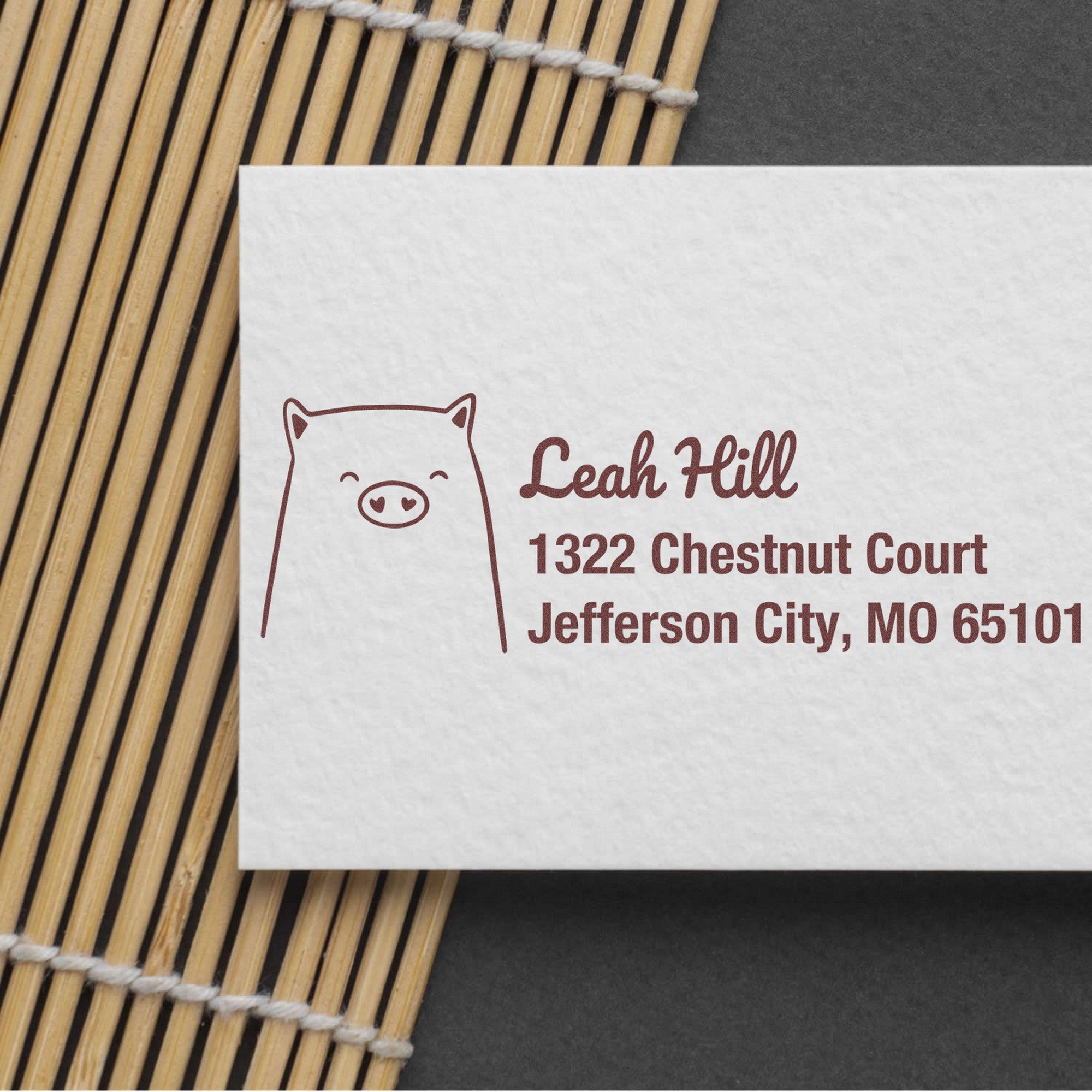 Slim Pre-Inked Cheery Pig Customized New Address Stamp on a white card with "Leah Hill, 1322 Chestnut Court, Jefferson City, MO 65101" in brown ink. The card is placed on a bamboo mat, showcasing the Customized Mailing Stamper design.
