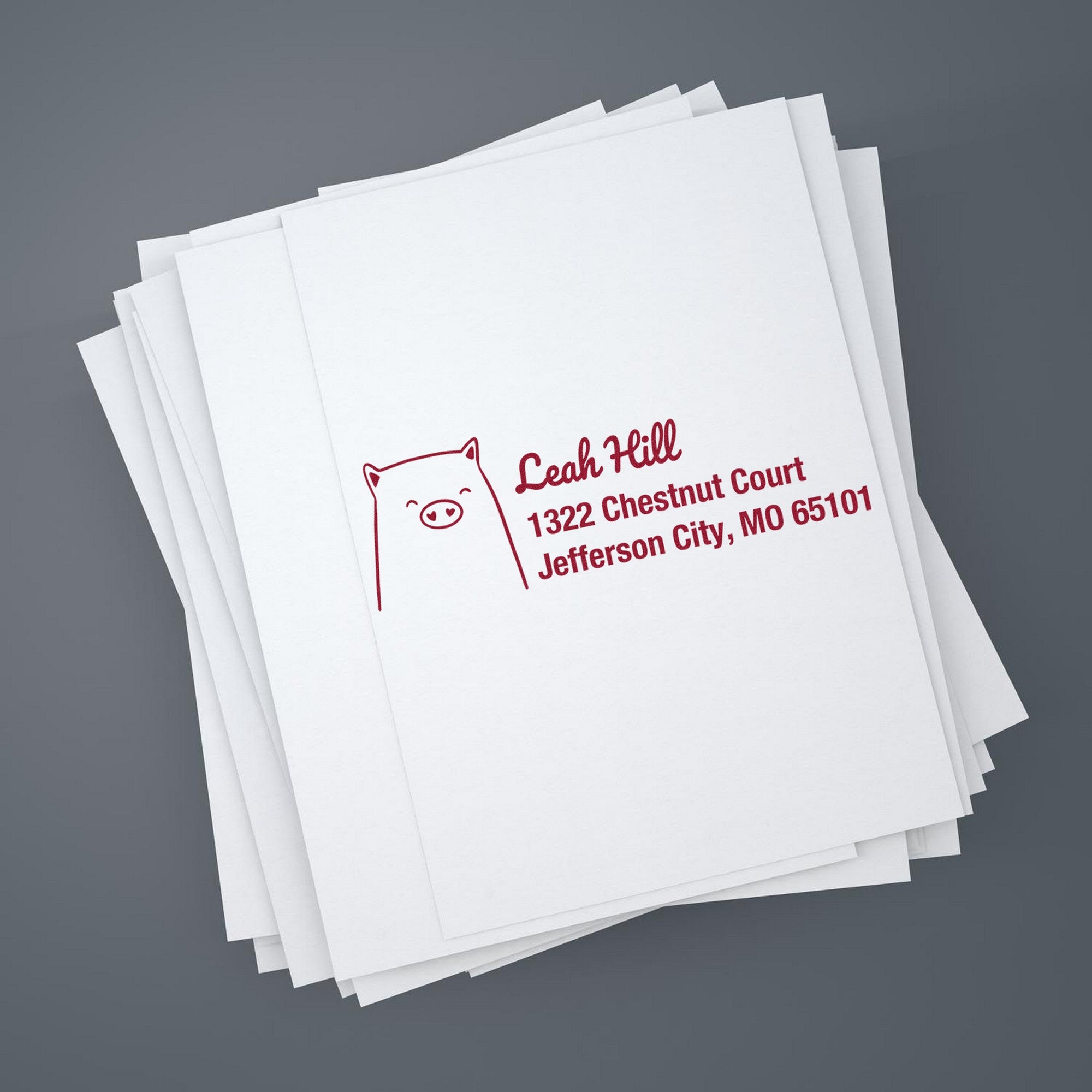 A stack of white papers featuring a Slim Pre-Inked Cheery Pig Customized New Address Stamp with red ink. The design includes a pig illustration and text for a customized return mail stamp, displaying an address in Jefferson City, MO.