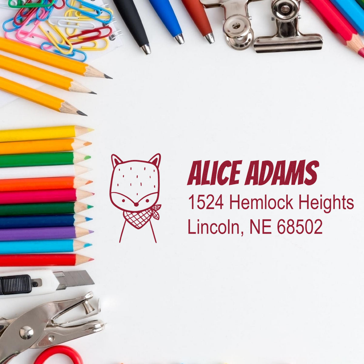 Slim Pre-Inked Fancy Fox Customized Address Label Stamp on white paper with colorful pencils, paperclips, and office supplies. Text reads "Alice Adams, 1524 Hemlock Heights, Lincoln, NE 68502." Ideal for Custom Mailing Stamper use.