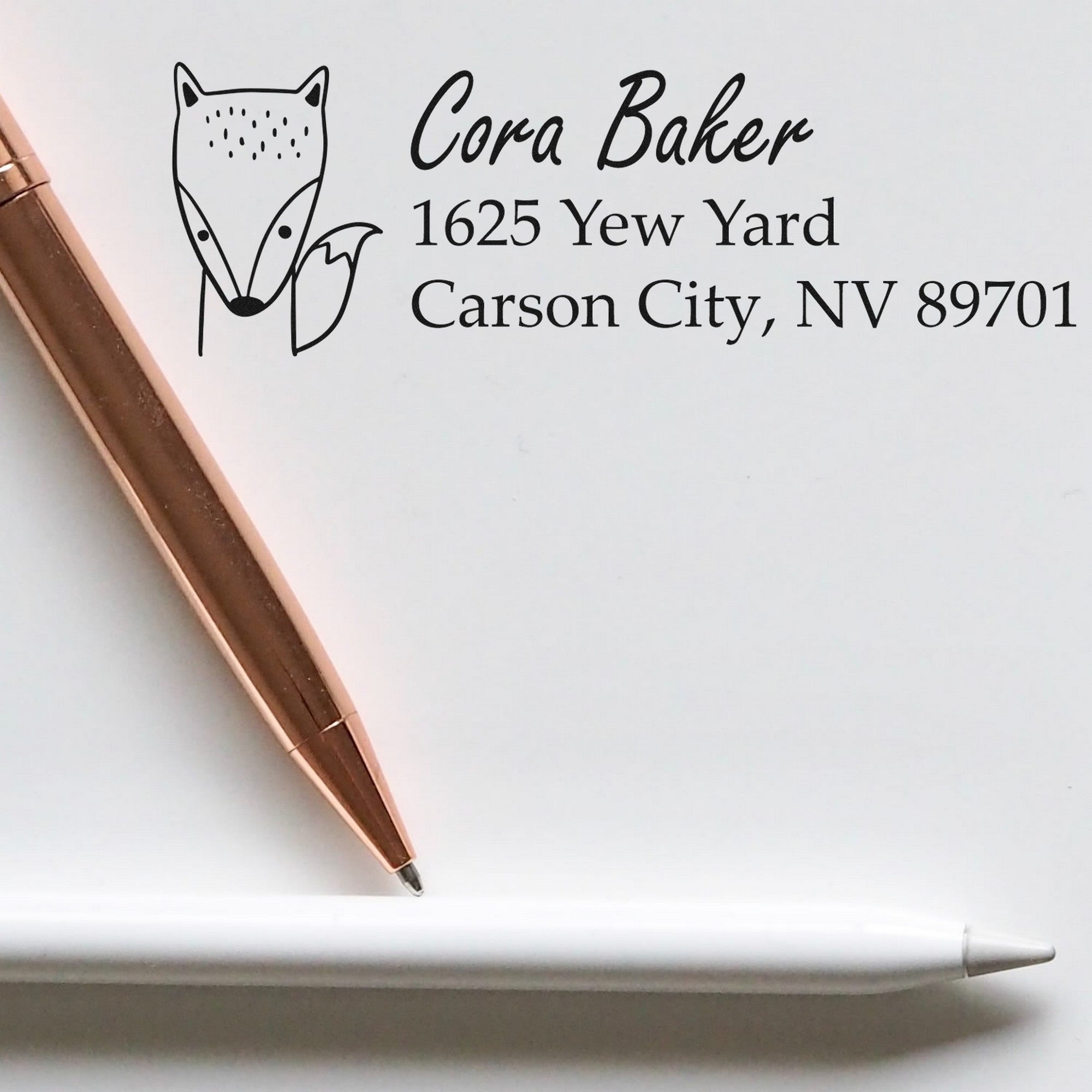 A personalized address stamp with a wood handle fast fox design is shown on paper. The stamp displays the name "Cora Baker" and an address in Carson City, NV. A rose gold pen and a white pen are placed beside the stamped text.
