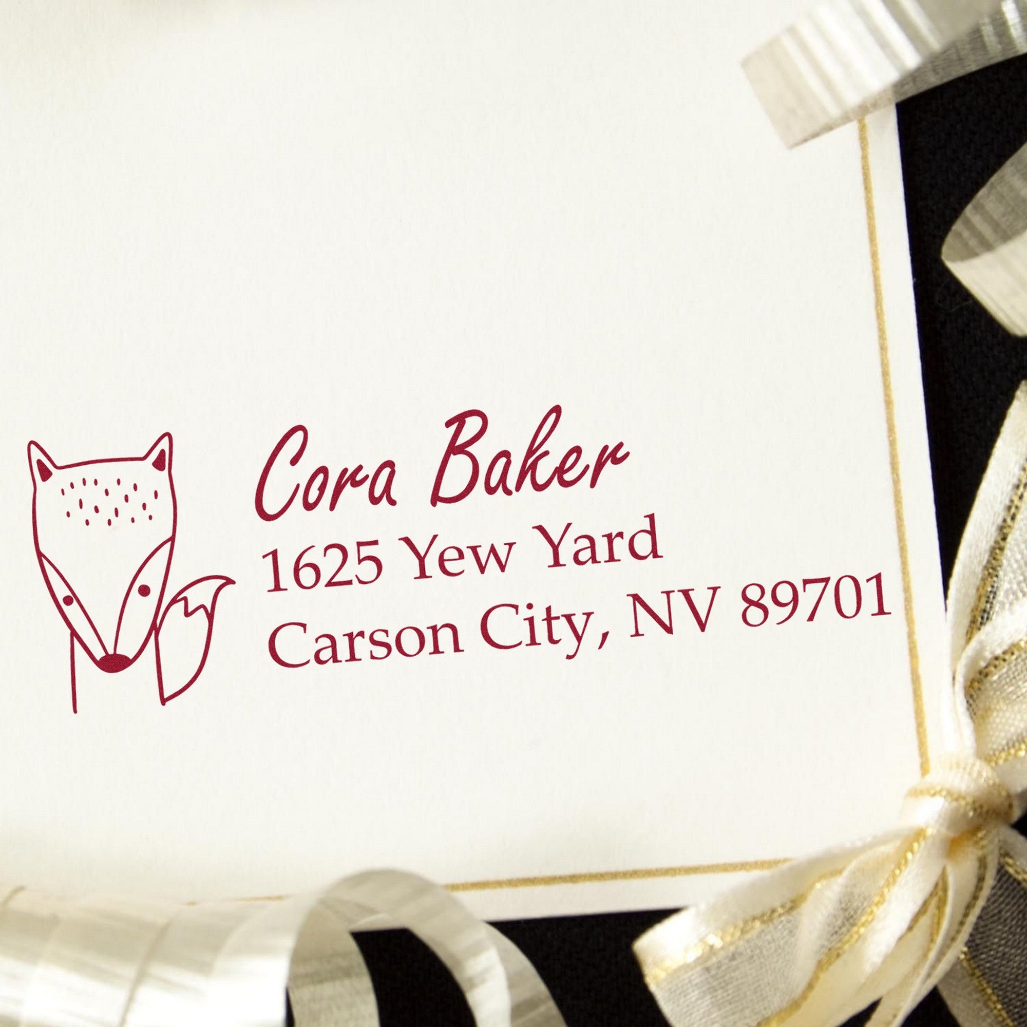 Image of a personalized return mail stamper with the text "Cora Baker, 1625 Yew Yard, Carson City, NV 89701" in red ink. The design features a fox illustration, showcasing the PSI Fast Fox Customize New Address Pre-Inked Stamp on a cream card with gold ribbon.