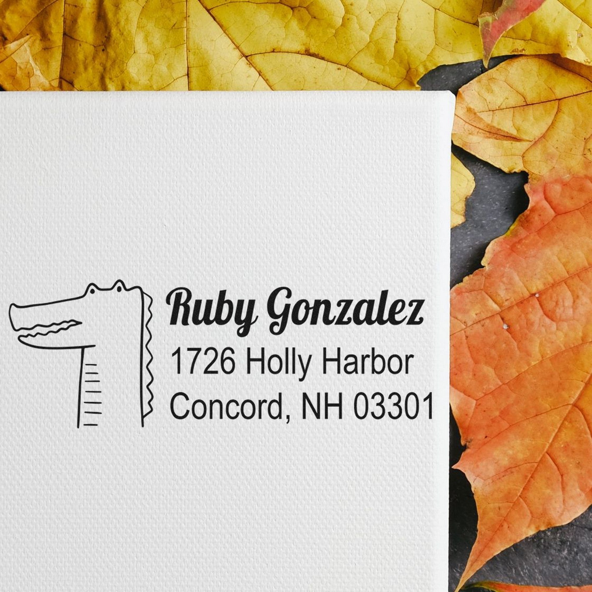 PSI Bright Eyed Crocodile Customize Home Address For Envelopes Pre-Inked Stamp displayed on a white canvas with autumn leaves. The Custom-Made Mailing Stamper features a crocodile design and personalized address for Ruby Gonzalez, 1726 Holly Harbor, Concord, NH 03301.