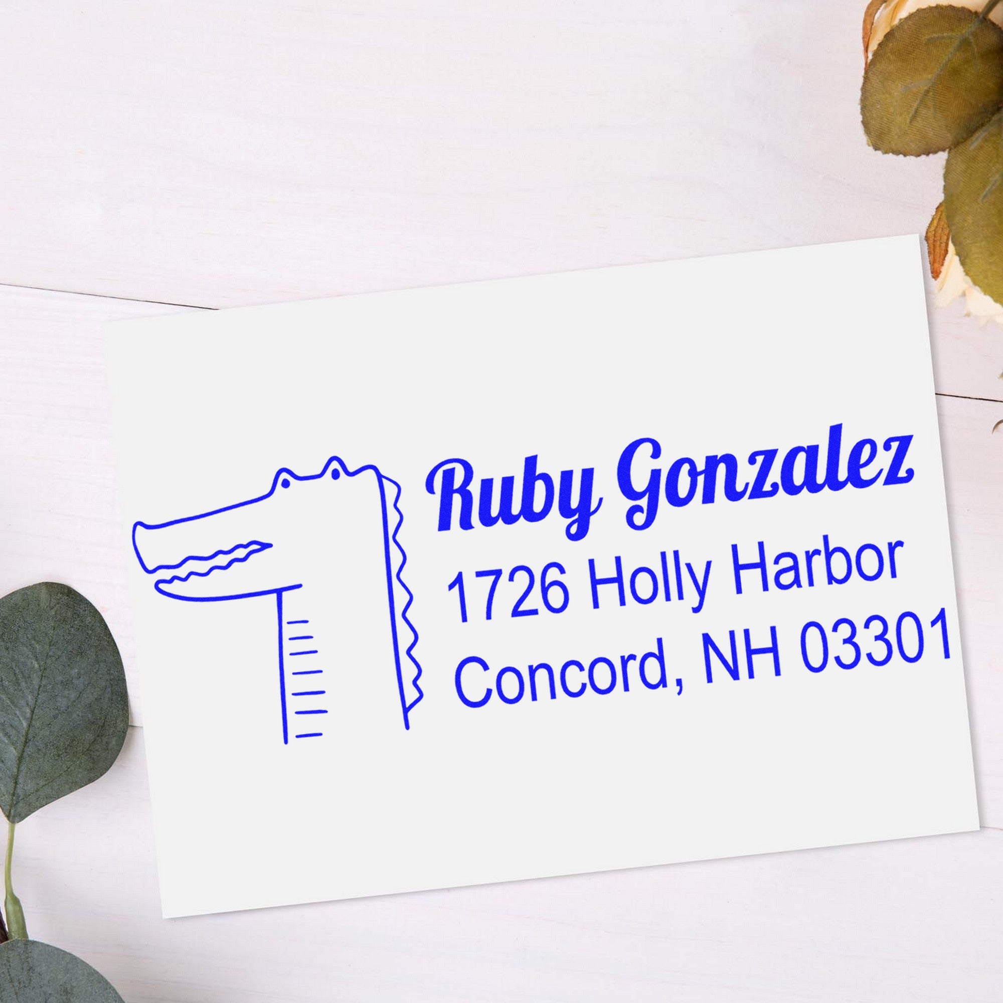 Wood handle bright-eyed crocodile customized mail rubber stamp on an envelope with personalized return mail stamper design, featuring the name Ruby Gonzalez and address in blue ink, surrounded by leaves on a light background.