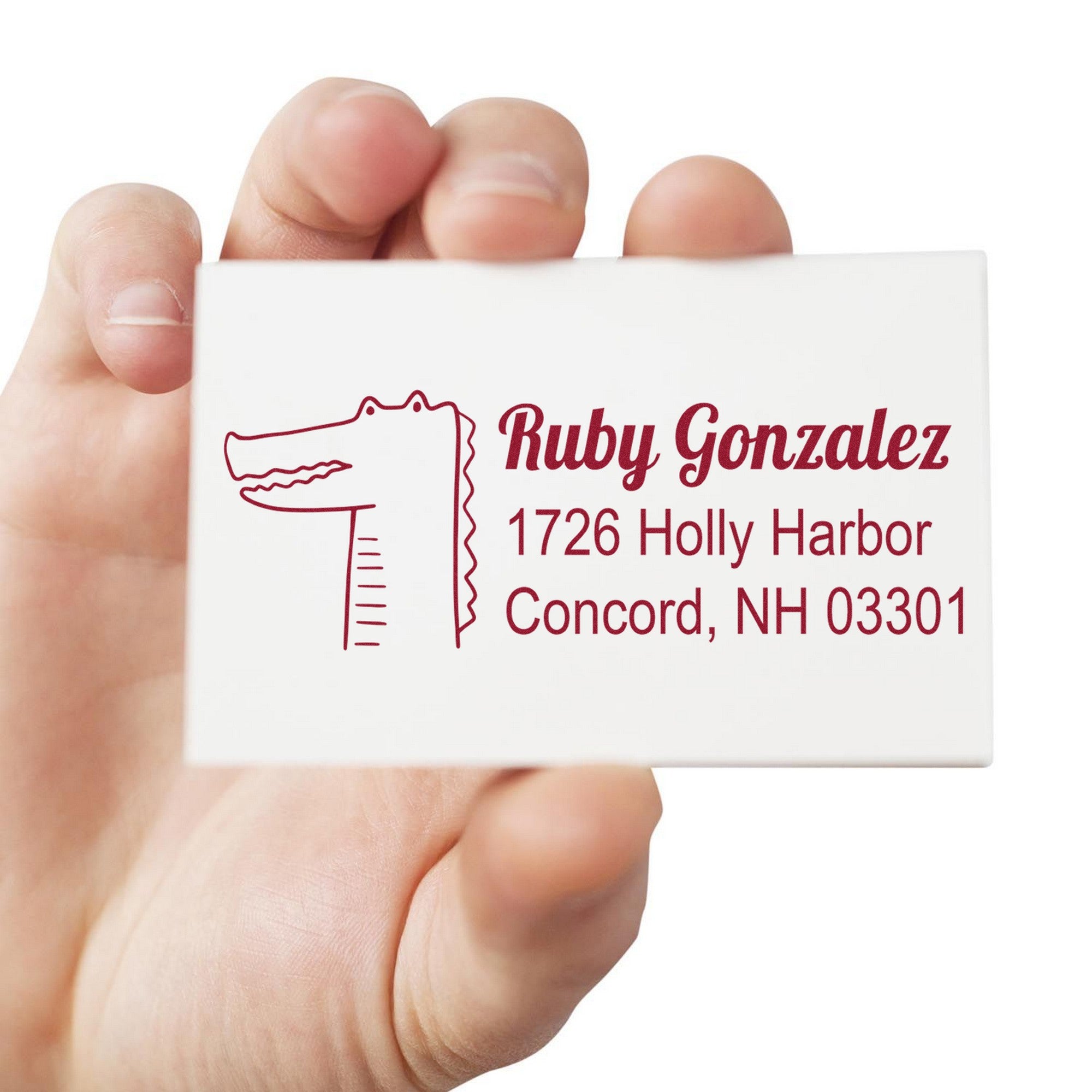 Hand holding a card with a red stamp design featuring a bright-eyed crocodile and text "Ruby Gonzalez, 1726 Holly Harbor, Concord, NH 03301." The stamp has a wood handle, labeled as a customized mail rubber stamp and custom address stamper.