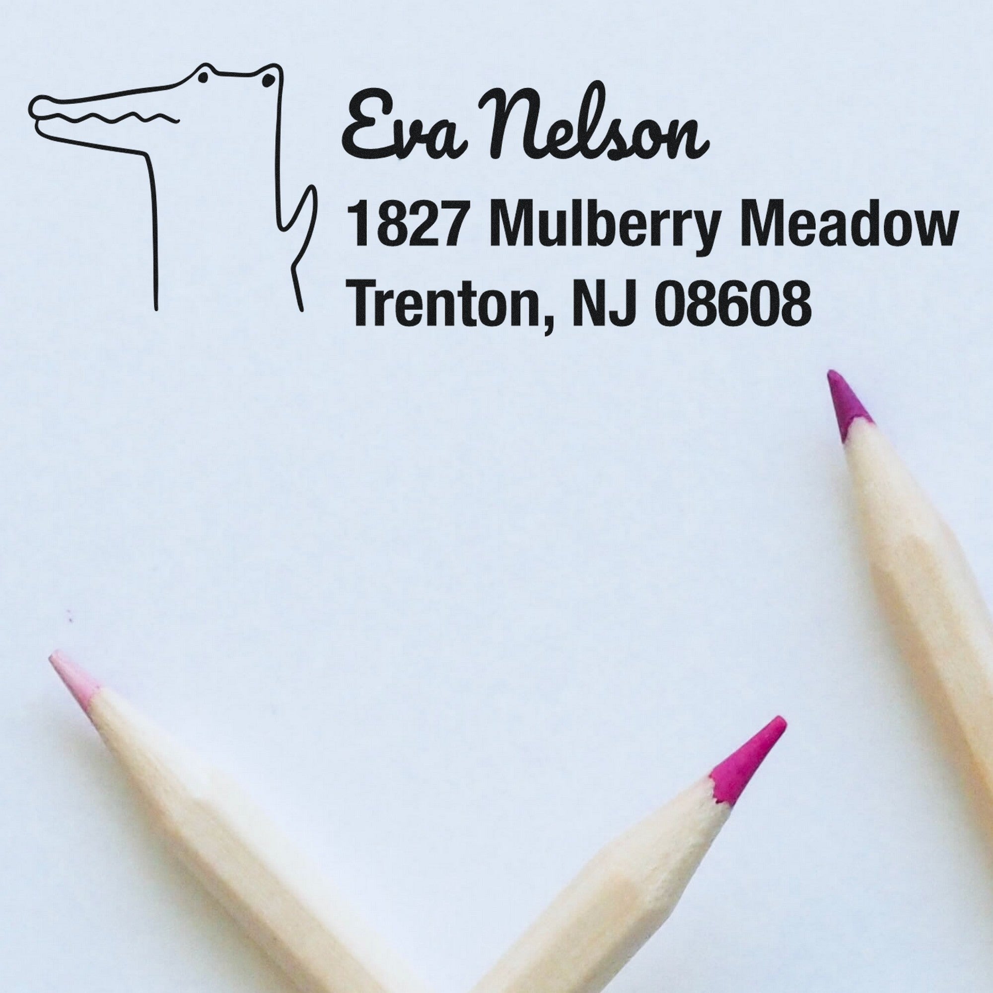 A Wood Handle Charming Crocodile Customized Home Address Rubber Stamp imprint on paper, showing Eva Nelson, 1827 Mulberry Meadow, Trenton, NJ 08608. Two pink-tipped pencils are nearby. This Customized Return Mail Stamper adds a playful touch to correspondence.
