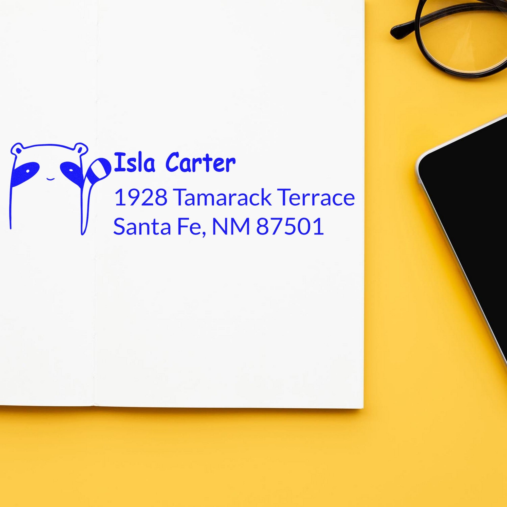 PSI Renegade Racoon Customizable Address Pre-Inked Stamp on a white card with blue text reading "Isla Carter, 1928 Tamarack Terrace, Santa Fe, NM 87501." Custom New Address Stamp design with a raccoon graphic, placed on a yellow surface next to a smartphone and glasses.