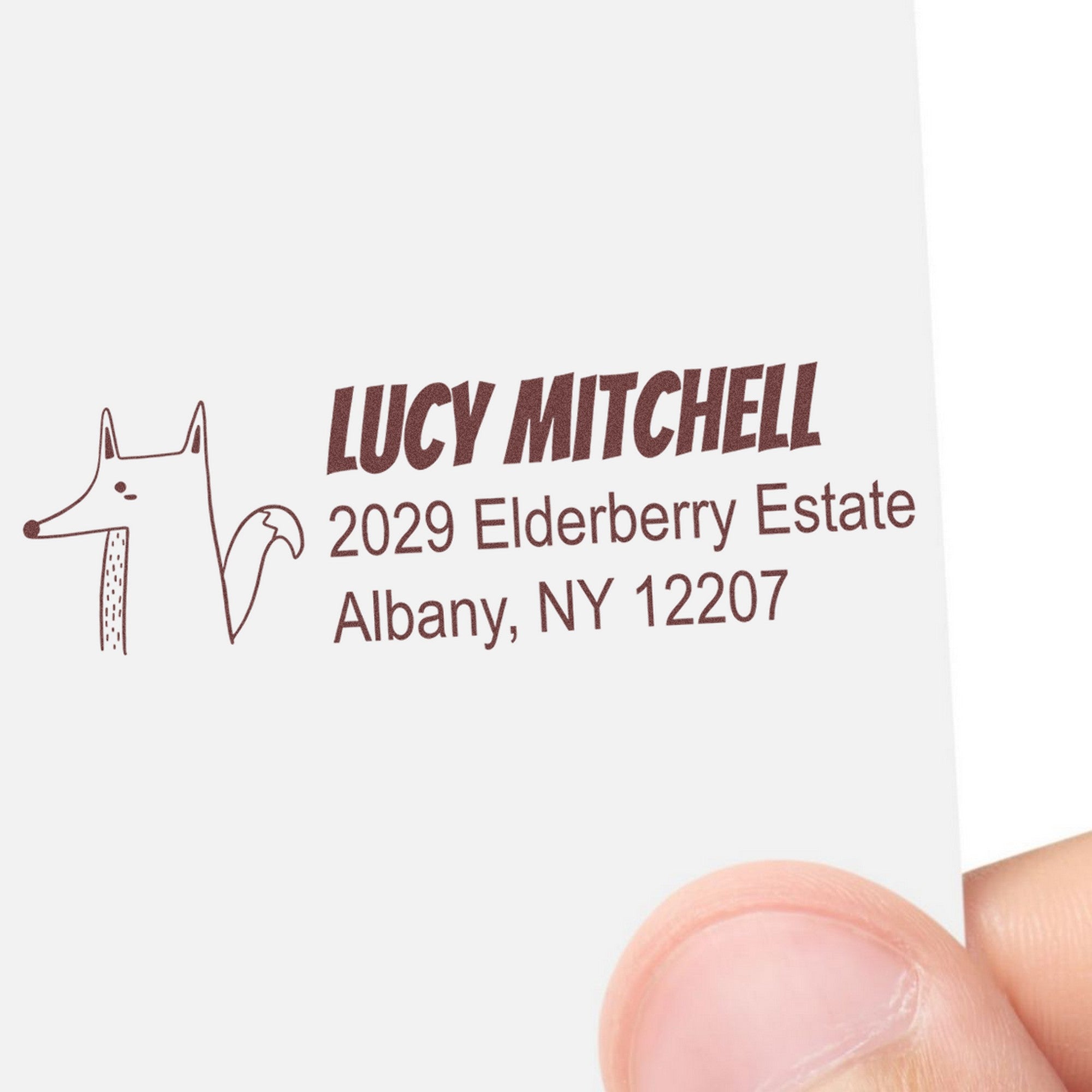 Image of a PSI Finicky Fox Customizable Return Address Pre-Inked Stamp imprint on paper, featuring a fox illustration and text: "Lucy Mitchell, 2029 Elderberry Estate, Albany, NY 12207." This custom-made new address stamp is held by a hand.
