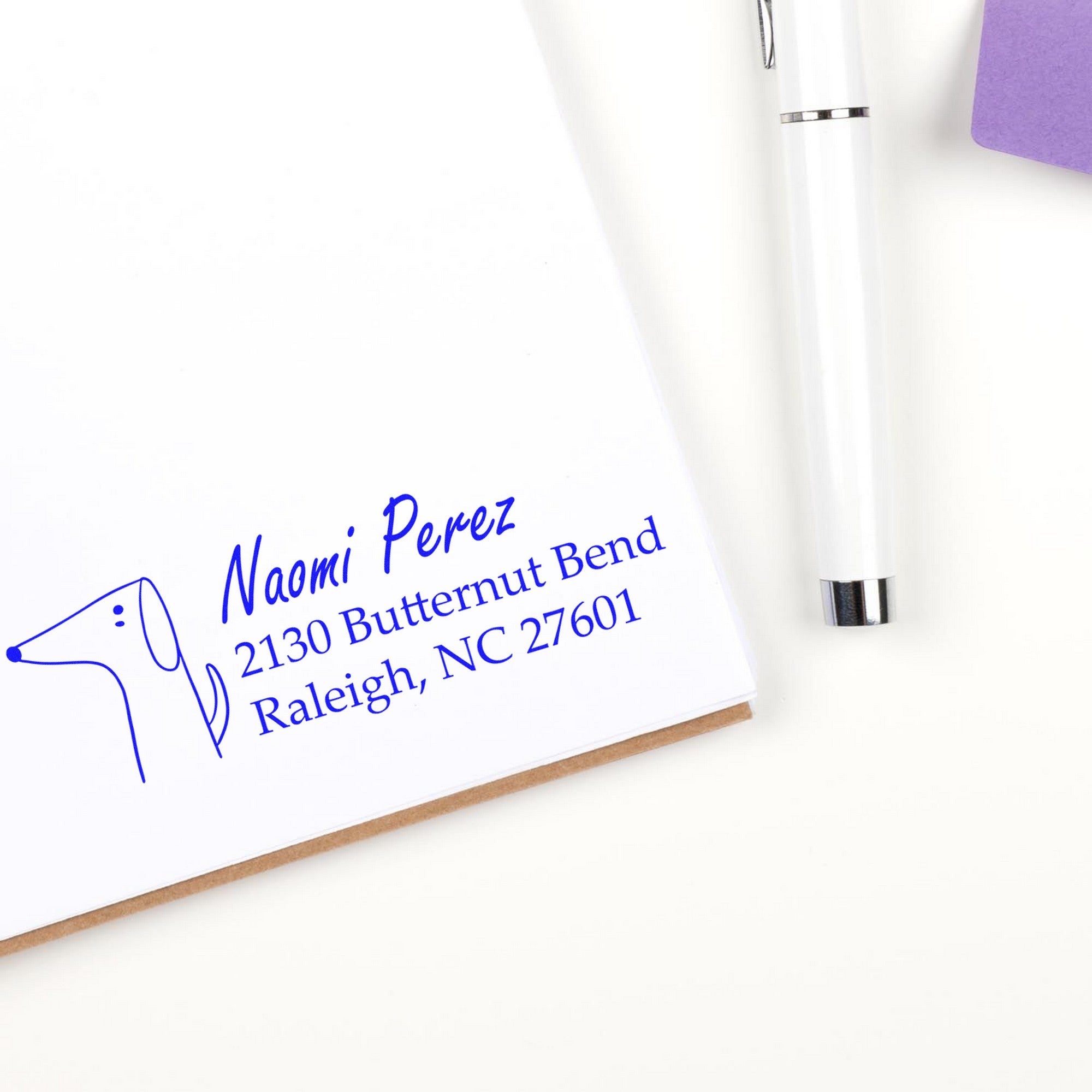 Delightful Dog Personalized Address Label Self-Inking Stamp on white paper, featuring a simple dog illustration and text: "Naomi Perez, 2130 Butternut Bend, Raleigh, NC 27601." A pen and purple notepad are nearby. Ideal for a personalized mailing stamp.