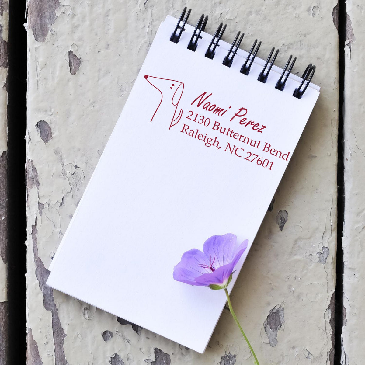 Slim Pre-Inked Delightful Dog Handmade Home Address Stamp on a notepad with a purple flower. The Customized New Address Stamper displays "Naomi Perez, 2130 Butternut Bend, Raleigh, NC 27601" in red ink, featuring a simple dog illustration.