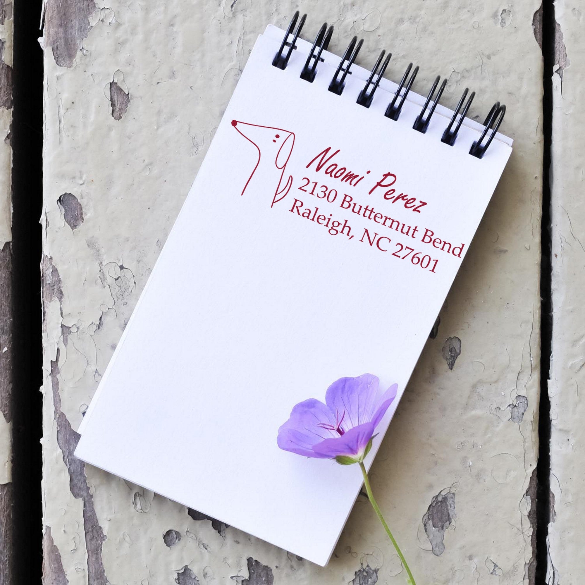 A notepad on a rustic surface features a PSI Delightful Dog Customizable Name and Address Pre-Inked Stamp with a dog illustration. The Custom New Address Stamp displays "Naomi Perez, 2130 Butternut Bend, Raleigh, NC 27601" in red ink, next to a purple flower.