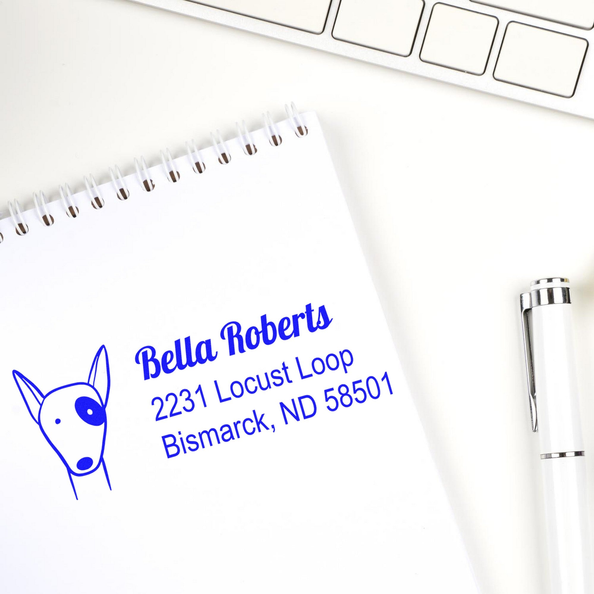 Wood Handle Perky Pooch Customized New Address Rubber Stamp used on a notepad displaying "Bella Roberts, 2231 Locust Loop, Bismarck, ND 58501" in blue ink. A Custom-Made Home Stamper is shown beside a pen and keyboard.