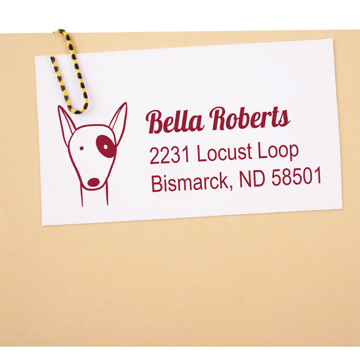 A beige envelope with a white card featuring a red dog illustration and text: "Bella Roberts, 2231 Locust Loop, Bismarck, ND 58501." The card is clipped with a black and yellow string. Keywords: Slim Pre-Inked Perky Pooch Handmade Address Return Stamp, Personalized New Address Stamp.