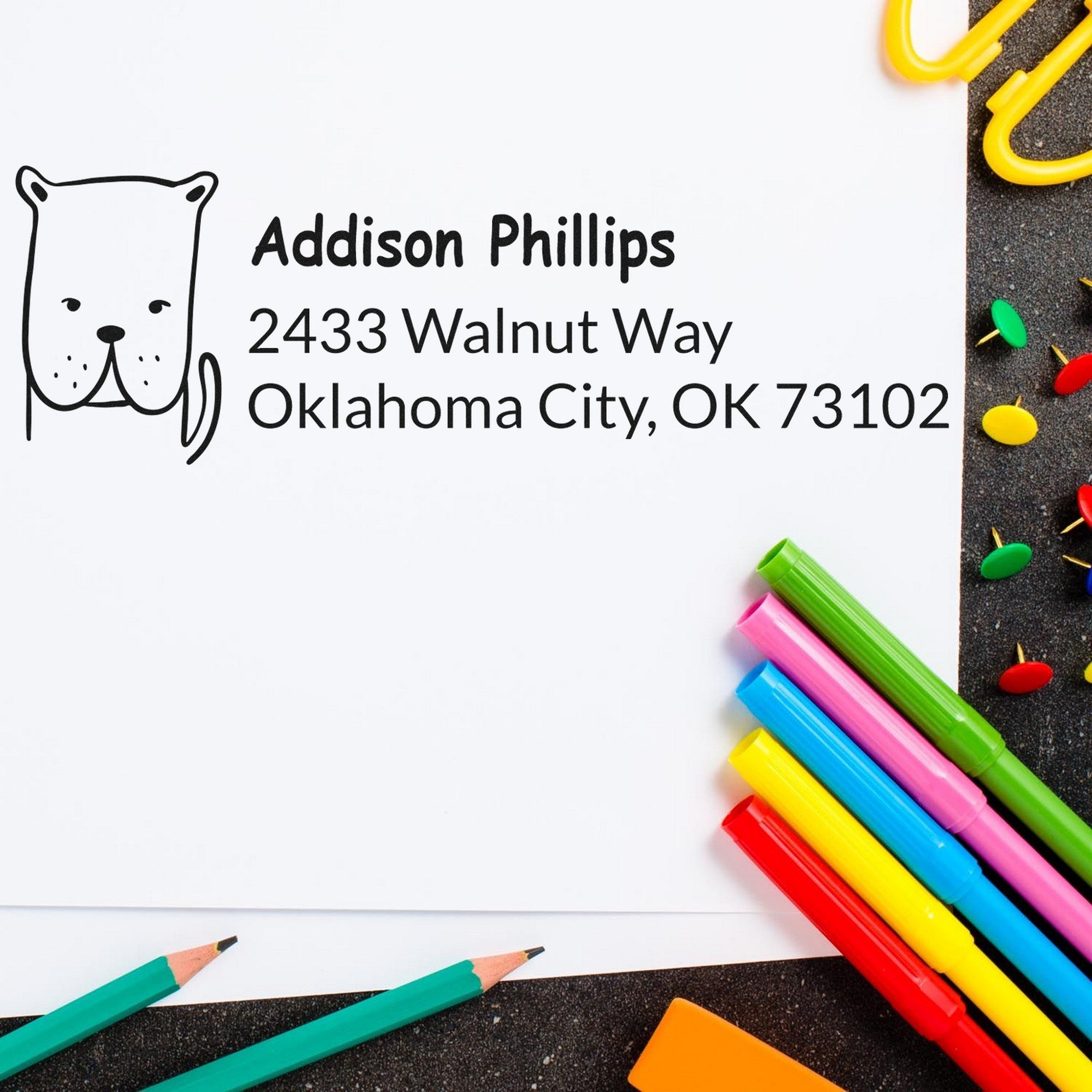 Daring Dog Custom Name and Address Self-Inking Stamp on white paper with "Addison Phillips, 2433 Walnut Way, Oklahoma City, OK 73102" text. Surrounded by colorful pens and paperclips. Customizable Home Stamp design.