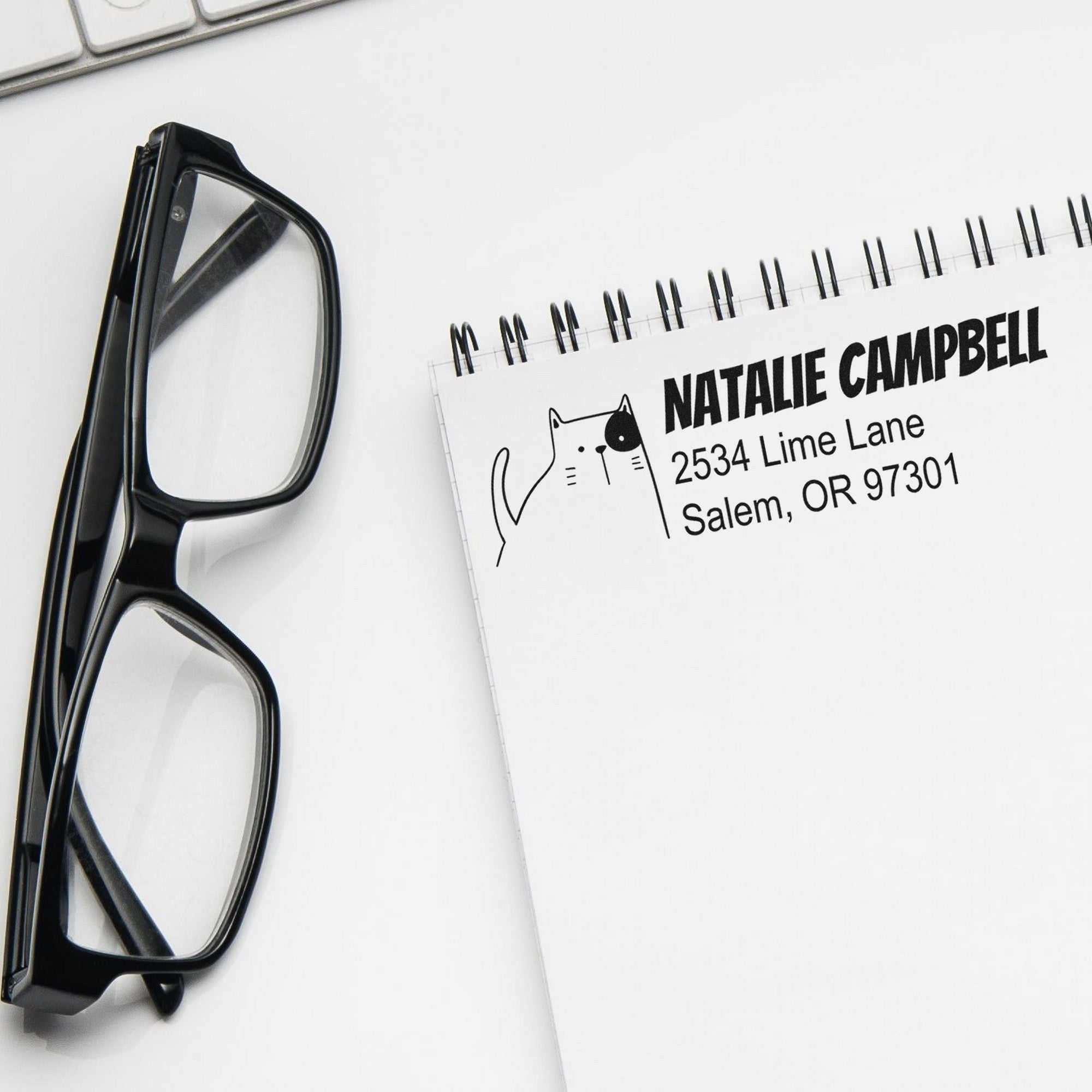 PSI Charming Cat Customizable Address Return Pre-Inked Stamp on a notepad with "Natalie Campbell, 2534 Lime Lane, Salem, OR 97301" printed. Glasses and a keyboard are nearby. Customizable New Address Stamper design features a cute cat illustration.