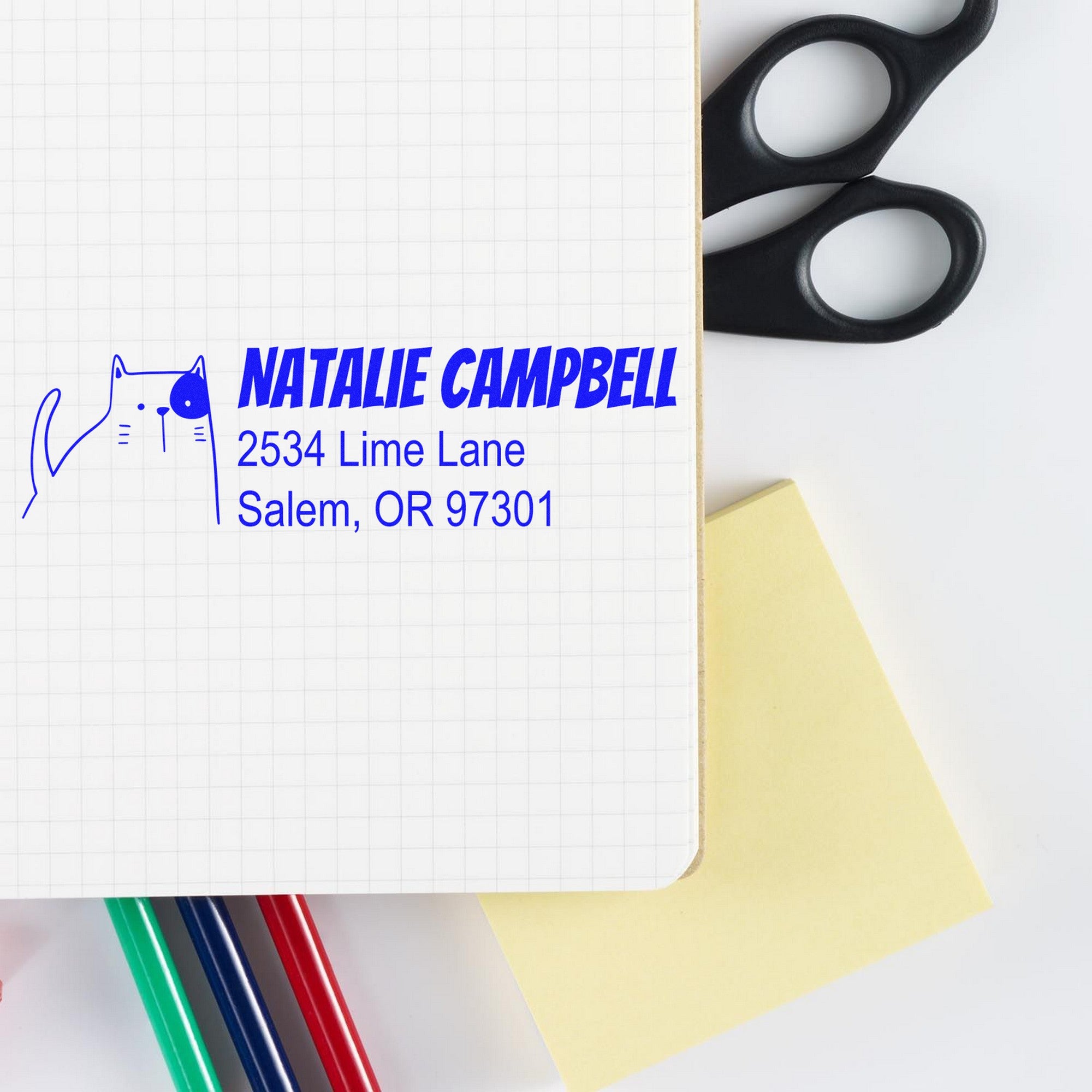 Image of a notebook with a PSI Charming Cat Customizable Address Return Pre-Inked Stamp featuring a cat design and the name "Natalie Campbell" with an address. Scissors, sticky notes, and colored pencils are nearby. Perfect for a Customizable Address Stamp.