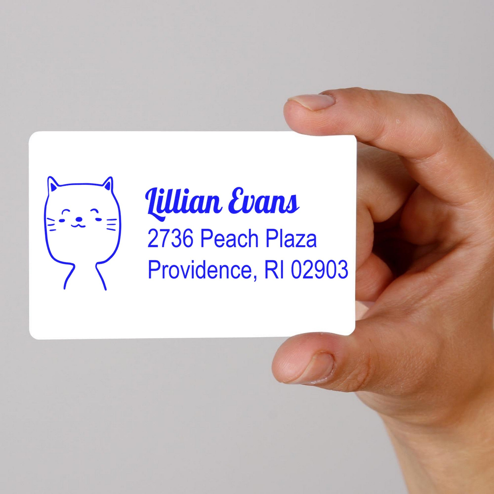 A hand holds a card stamped with a PSI Happy Cat Customizable Mail Address Pre-Inked Stamp, featuring a cute cat illustration and text. The Customized Home Stamper displays the name Lillian Evans and an address in Providence, RI.
