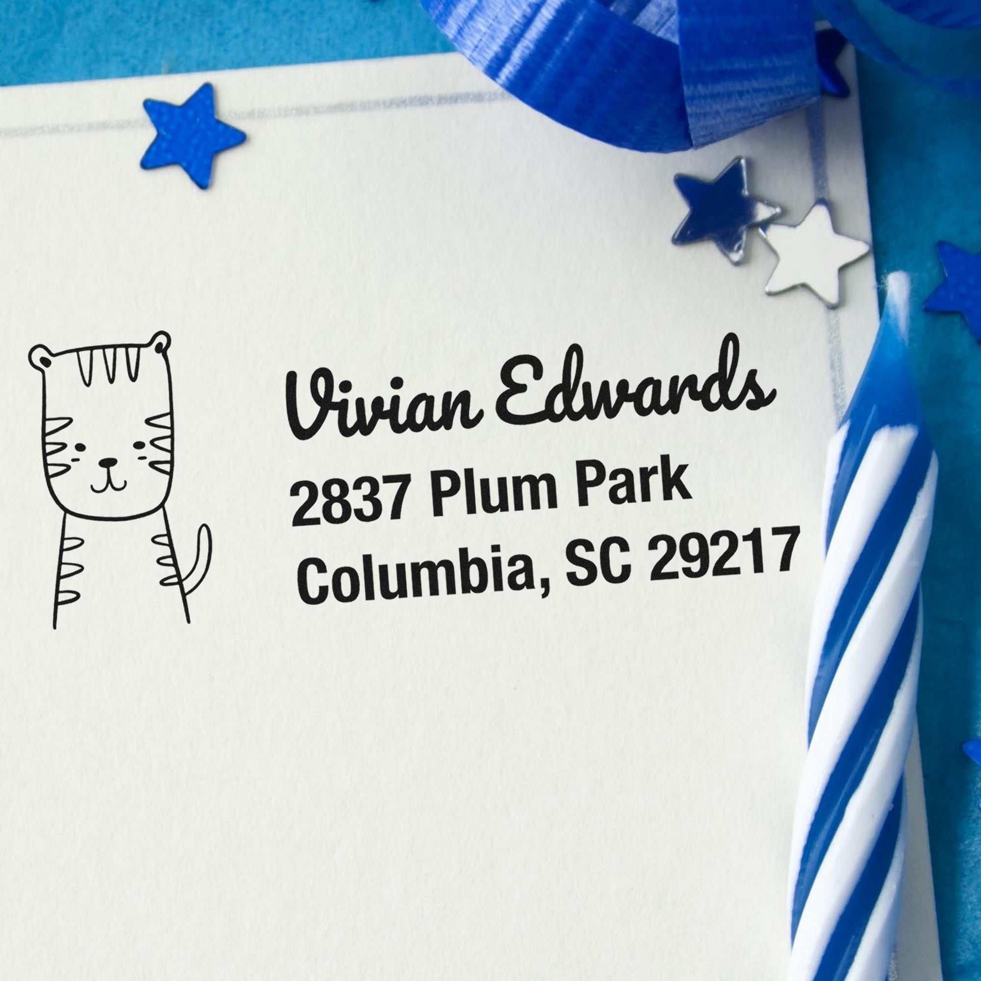 PSI Terrific Tiger Customizable New Address Pre-Inked Stamp on an envelope with a cute tiger design. Text reads "Vivian Edwards, 2837 Plum Park, Columbia, SC 29217." Blue stars and a striped candle are nearby. Custom-Made Return Mail Stamp.