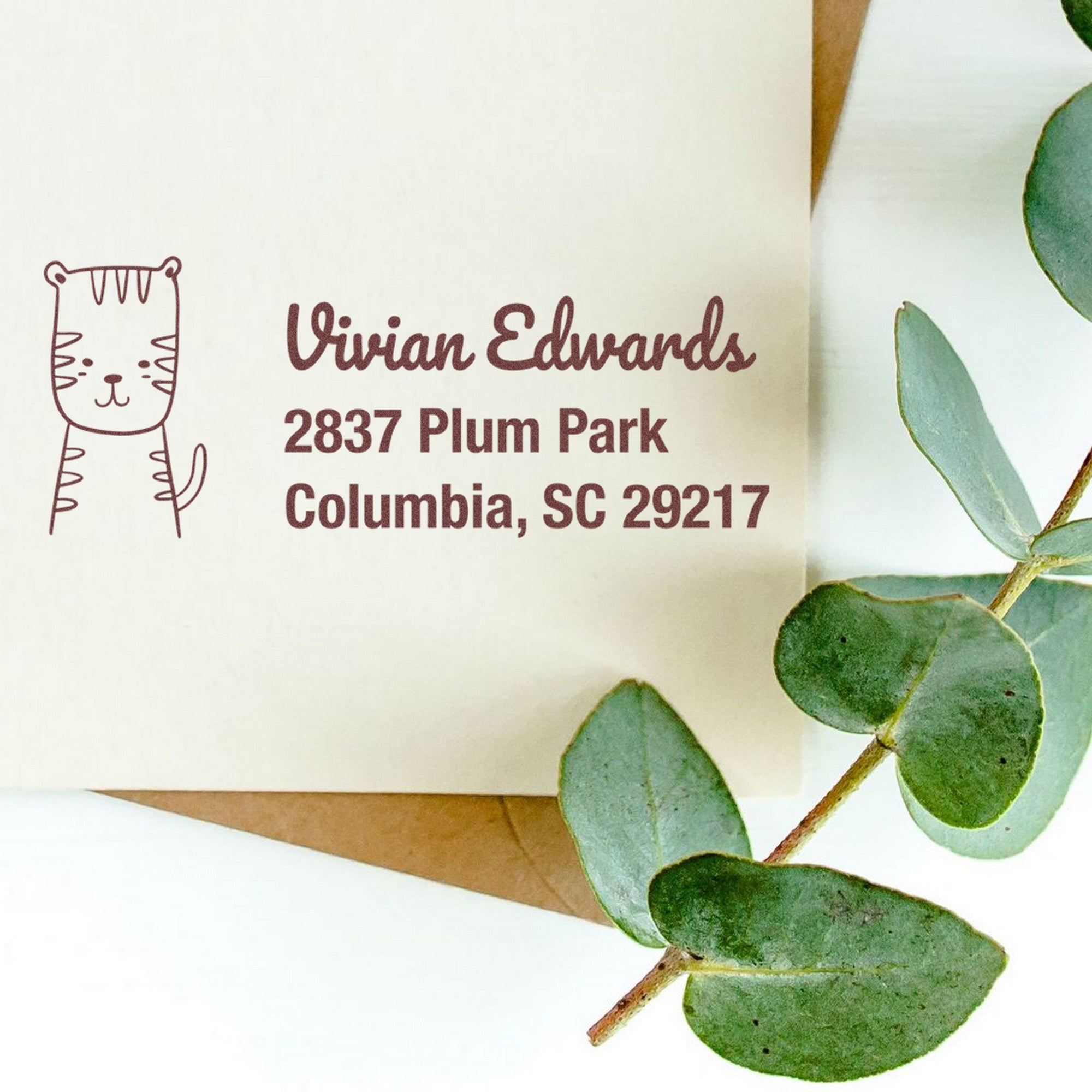 PSI Terrific Tiger Customizable New Address Pre-Inked Stamp on an envelope with a cute tiger illustration and the address "Vivian Edwards, 2837 Plum Park, Columbia, SC 29217." Eucalyptus leaves are placed beside the Custom Mailing Stamper.