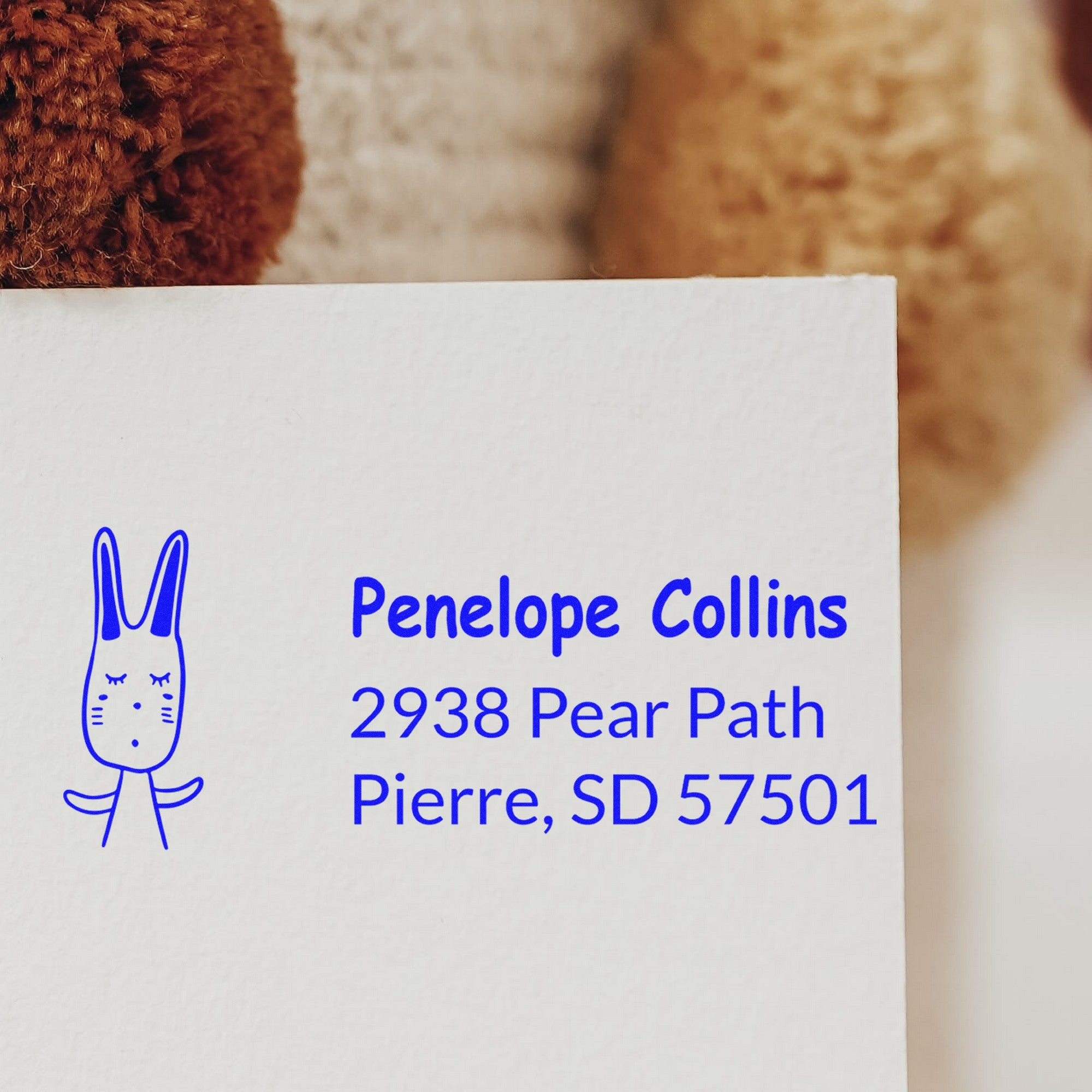Bouncy Bunny Custom Mailing Address Self-Inking Stamp on an envelope, featuring a cute bunny design. The Custom-Made New Address Stamper displays "Penelope Collins, 2938 Pear Path, Pierre, SD 57501" in blue ink.