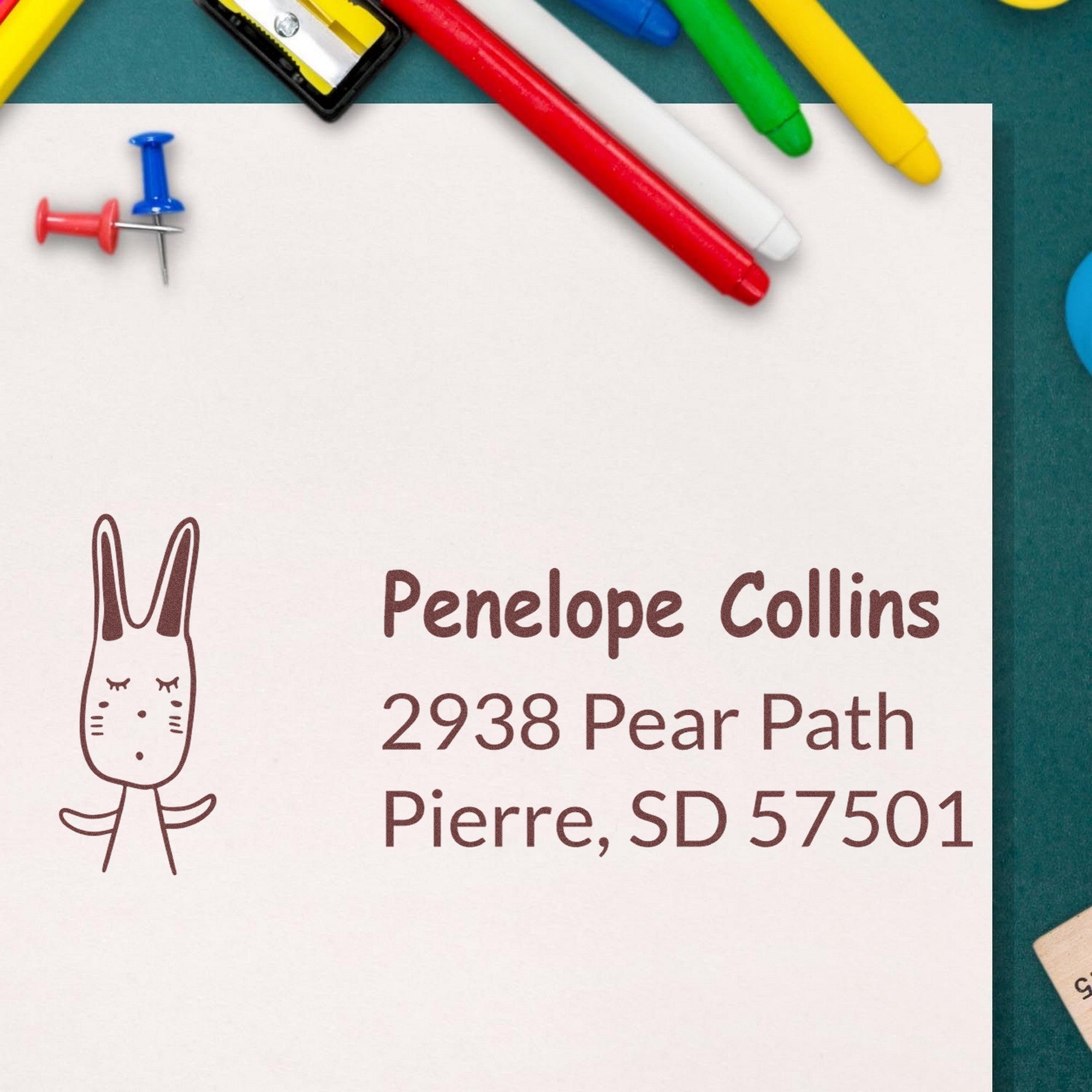 PSI Bouncy Bunny Customizable Home Address For Envelopes Pre-Inked Stamp on paper with the name Penelope Collins and address. Surrounding items include colorful pens and push pins. Customizable Address Stamp design features a cute bunny illustration.