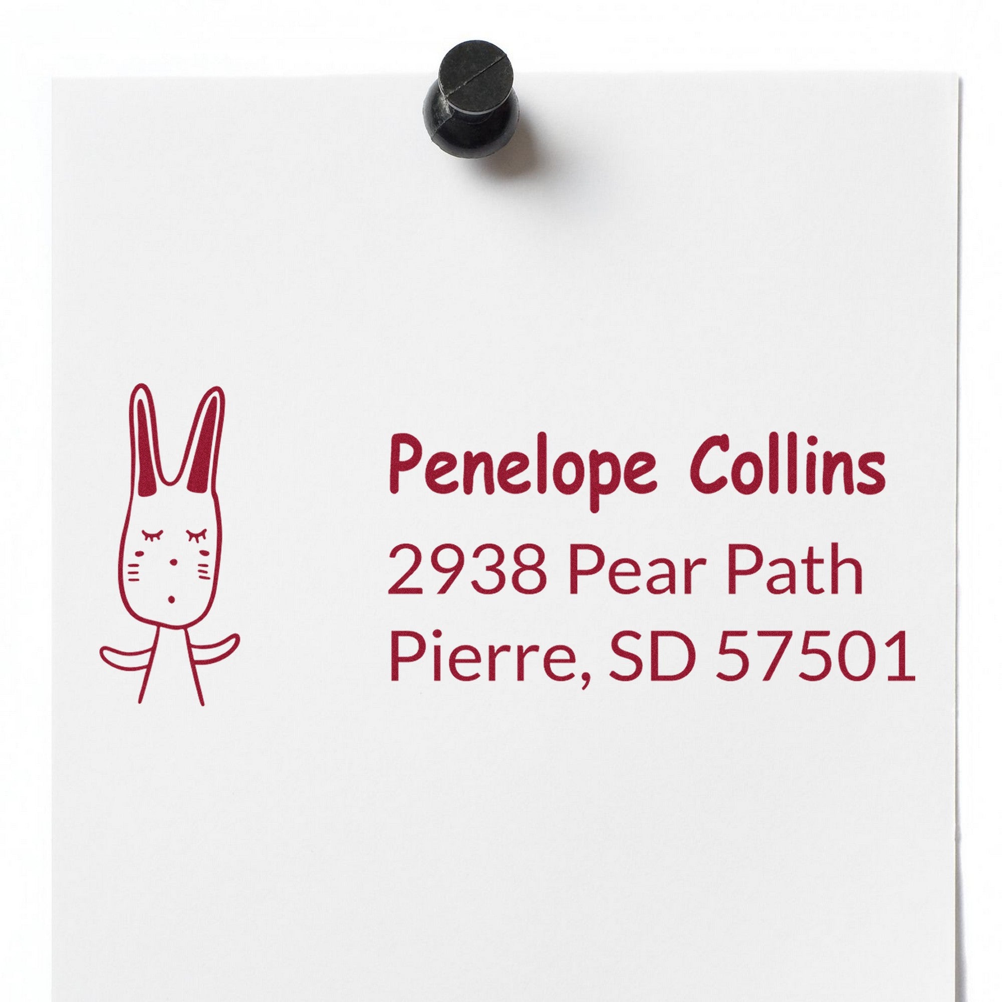 A white paper pinned to a board displays a PSI Bouncy Bunny Customizable Home Address For Envelopes Pre-Inked Stamp. The design features a cute bunny illustration and text reading "Penelope Collins, 2938 Pear Path, Pierre, SD 57501." Ideal for a Customizable New Address Stamper.