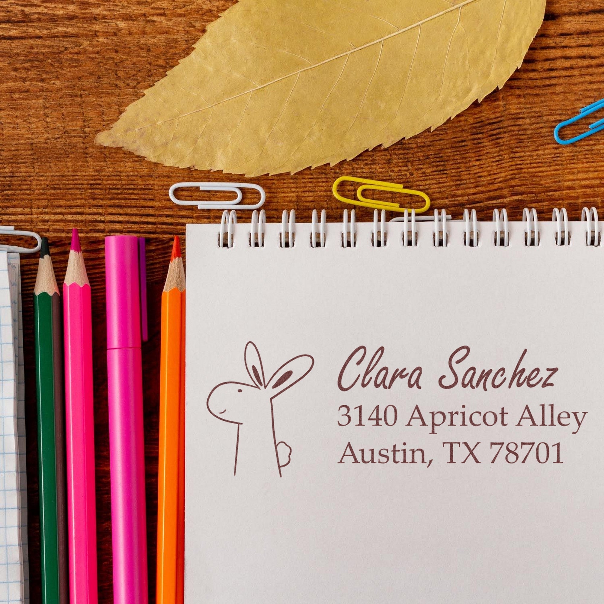 PSI Hopping Bunny Personalized Address Pre-Inked Stamp on a notepad with "Clara Sanchez, 3140 Apricot Alley, Austin, TX 78701" text. Custom Mailing Stamp design features a bunny illustration. Nearby are colored pencils, paperclips, and a leaf on a wooden surface.