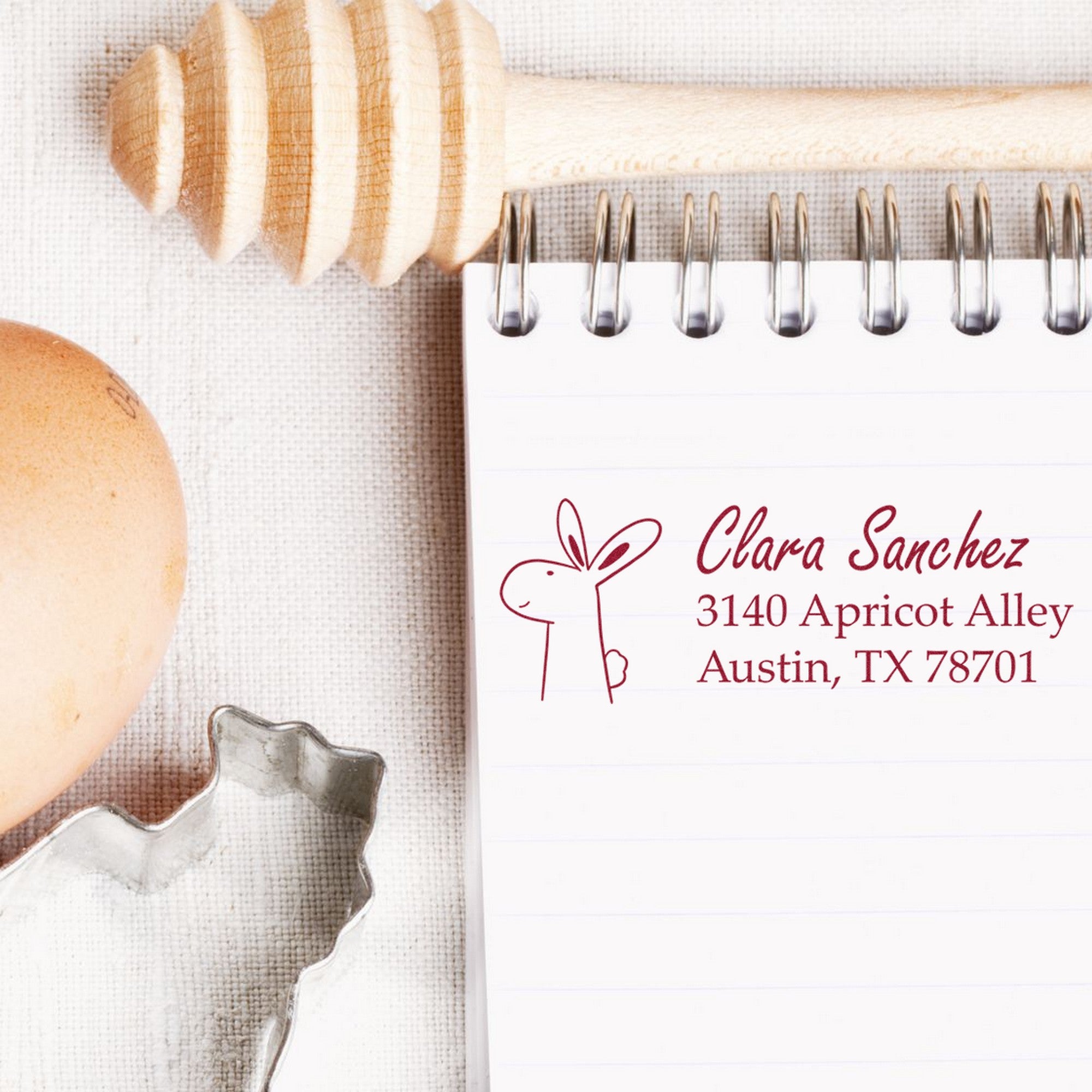Hopping Bunny Custom New Address Self-Inking Stamp on a notepad with "Clara Sanchez, 3140 Apricot Alley, Austin, TX 78701" in red ink. The Custom New Address Stamp is next to a honey dipper, egg, and leaf-shaped cookie cutter.