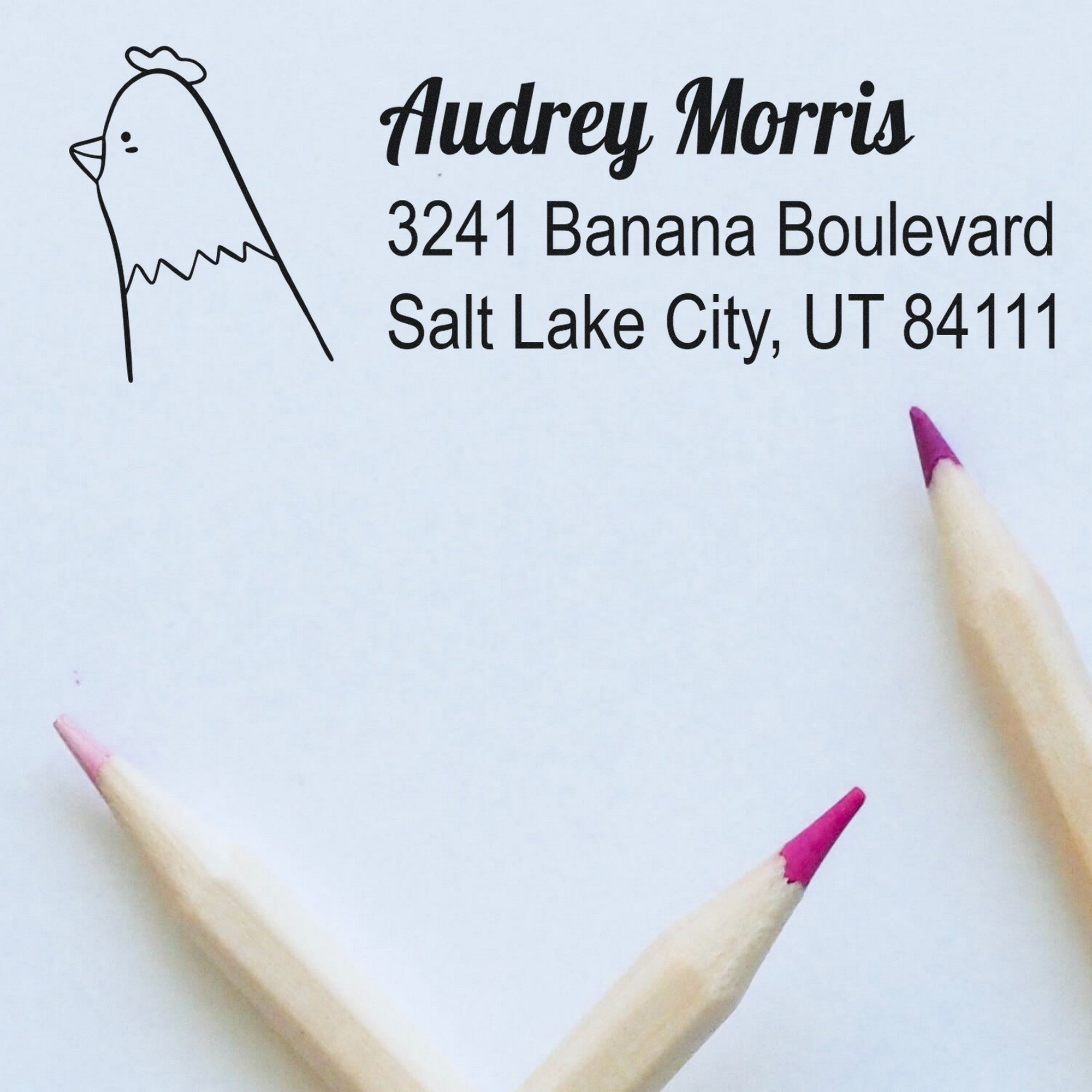 Wood handle clucking chicken handmade mailing address rubber stamp on paper, displaying "Audrey Morris, 3241 Banana Boulevard, Salt Lake City, UT 84111." Custom mailing stamper design with pink-tipped pencils nearby.