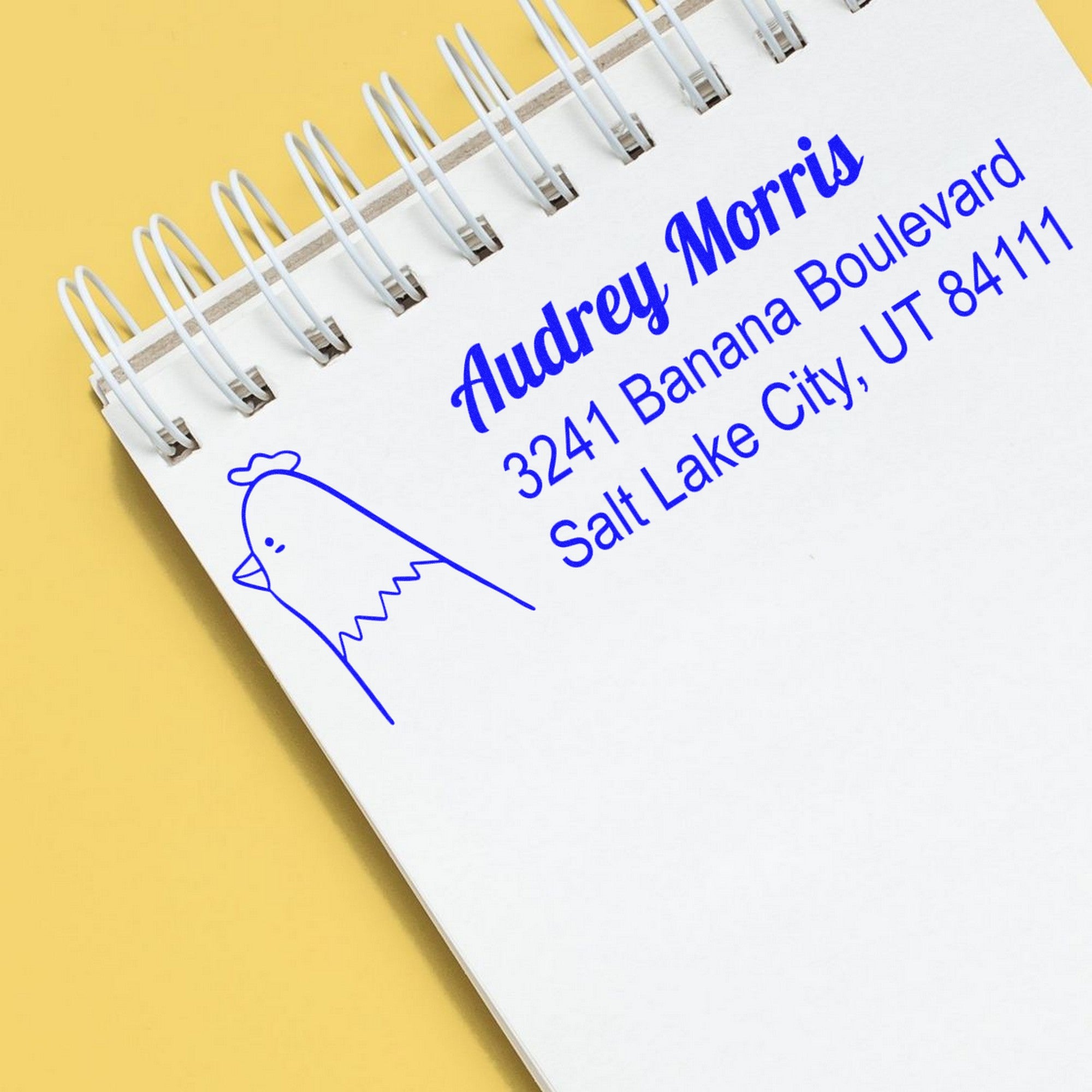 Wood handle clucking chicken handmade mailing address rubber stamp on a notepad. The custom-made new address stamp displays "Audrey Morris, 3241 Banana Boulevard, Salt Lake City, UT 84111" in blue ink, with a chicken illustration.